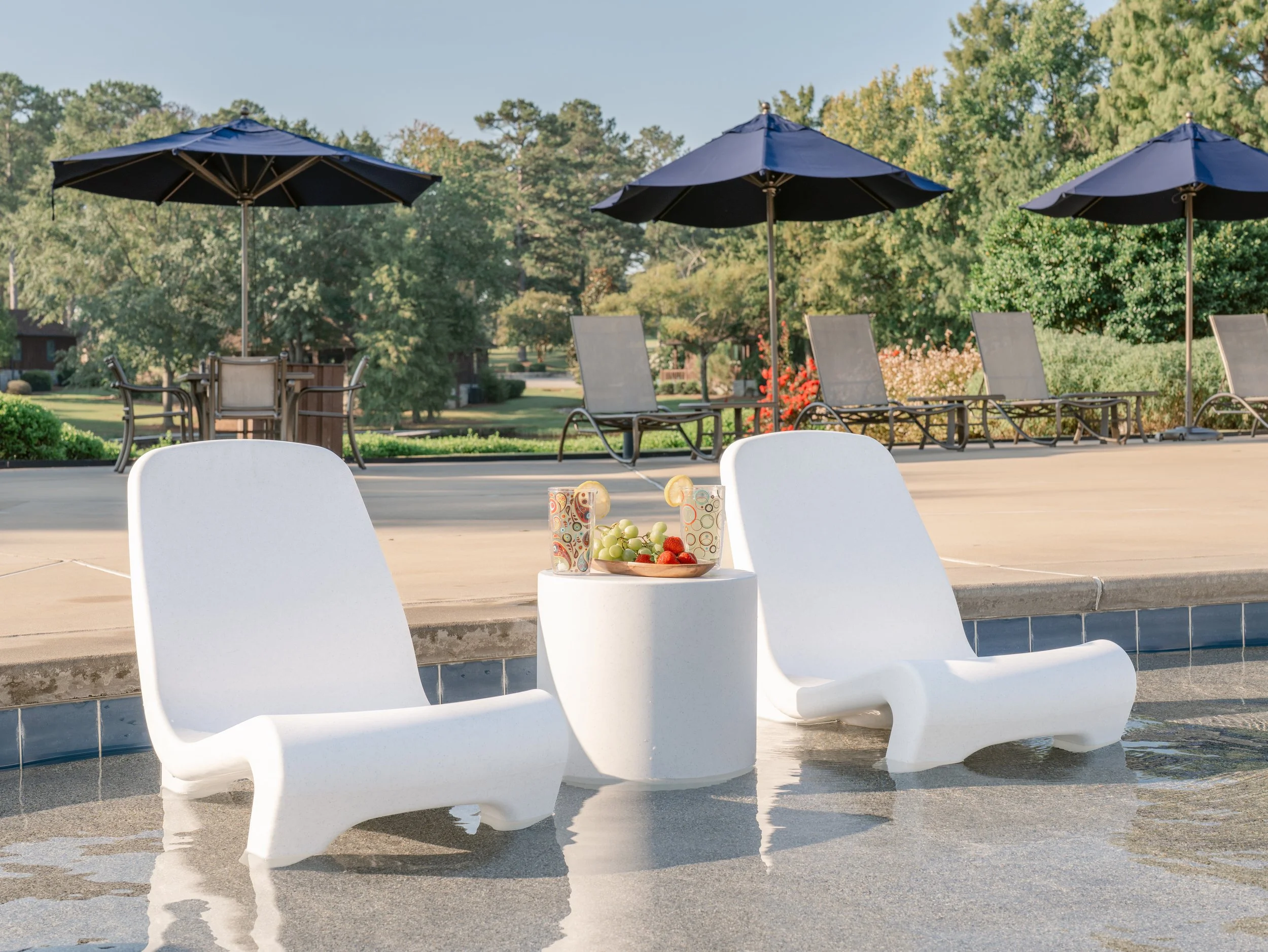 Two white lounge chairs in pool water with a small round table between them holding two glasses of fruit drinks, grapes, and strawberries. In the background, there are three black umbrellas over lounge chairs, surrounded by greenery and trees.