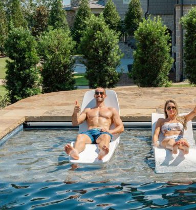 A man and a woman are relaxing on lounge chairs partially submerged in a swimming pool. The man is shirtless, wearing blue swim trunks, sunglasses, and smiling while giving a thumbs-up. The woman is wearing sunglasses and a colorful swimsuit, with he