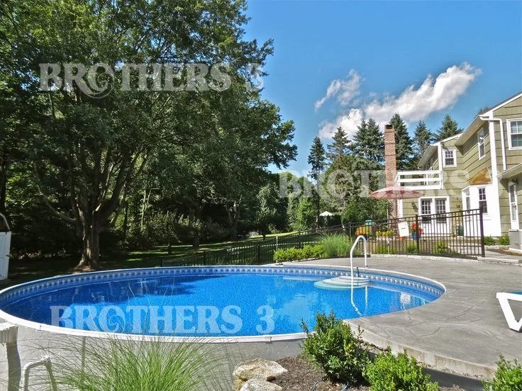 An outdoor backyard pool area with a clear blue swimming pool, surrounded by a concrete deck, with a house in the background, and trees and greenery around.