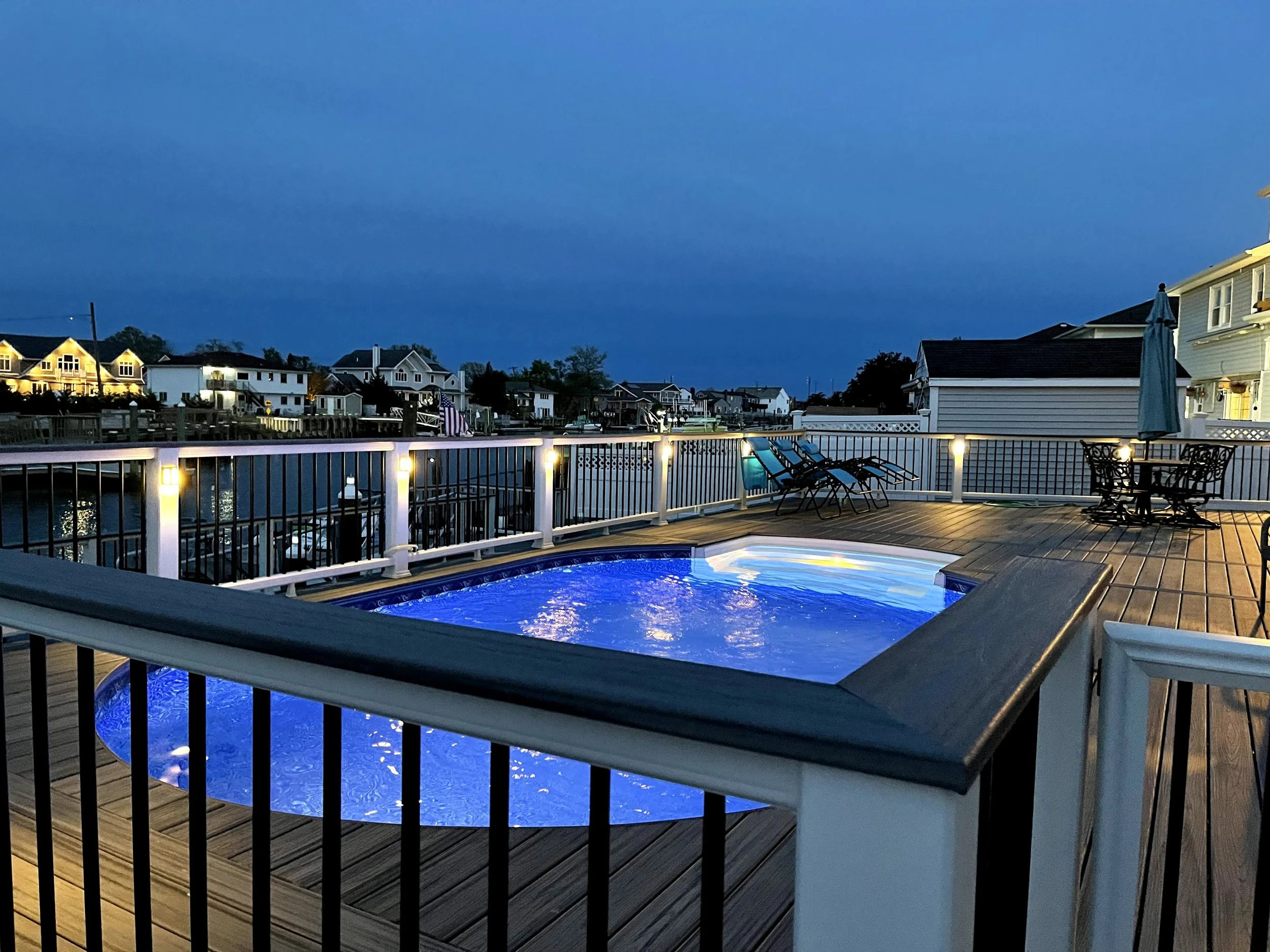 A backyard with a small lit swimming pool on a wooden deck, surrounded by a white railing, with house lights in the background during evening or early night.