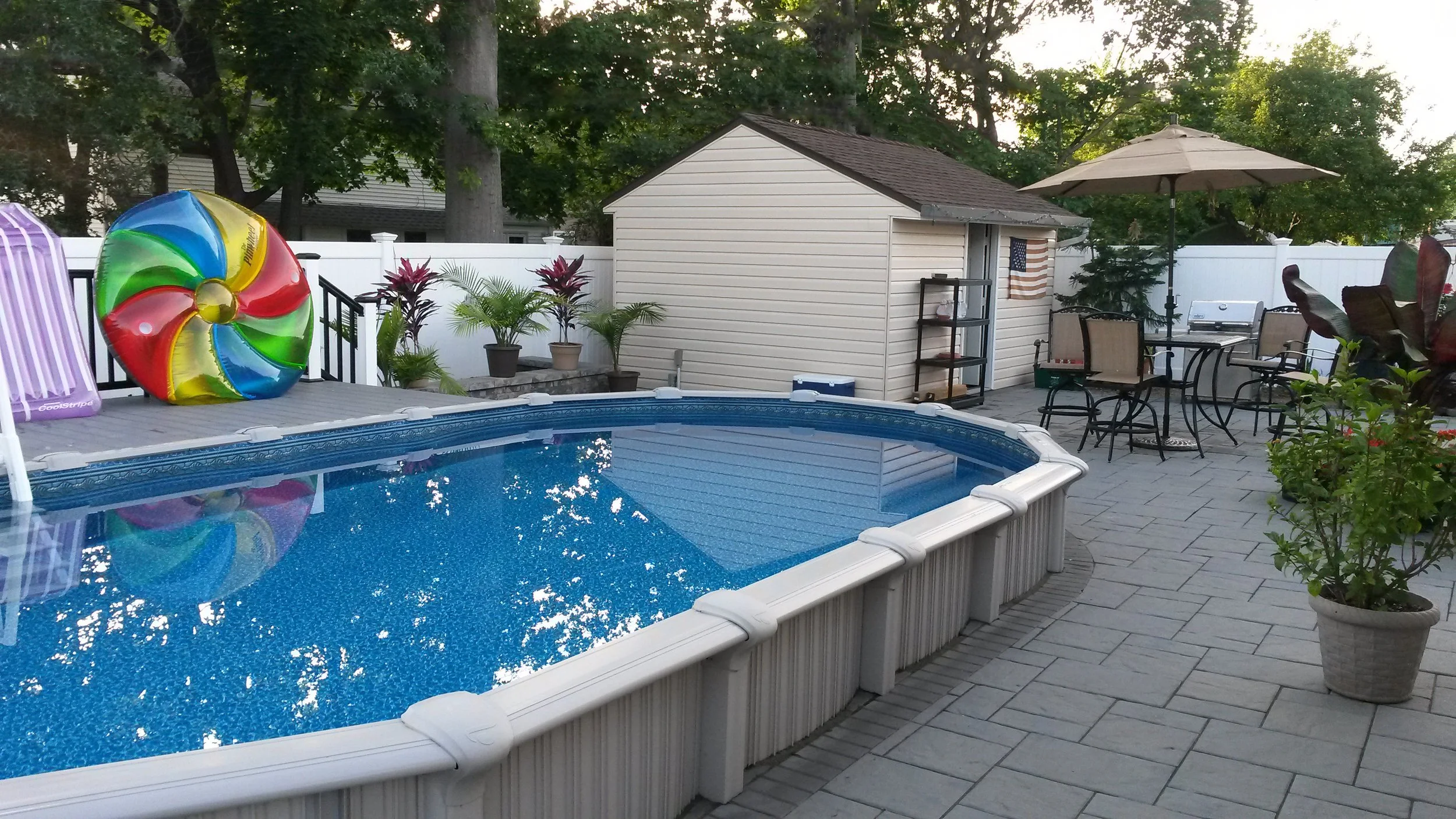 Backyard pool area with inflatable toy, lounge chair, potted plants, small shed, and outdoor patio furniture under an umbrella.