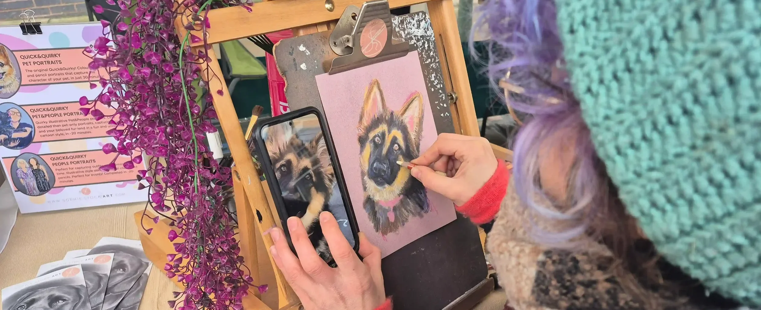 A cropped image of a lady sketching a german shepherd on a piece of pink paper, mounted on a clipboard, on an easel. There is a pink wisteria hanging to the left of the easel, and signage in the background shows 'Sophie Stock Art'.