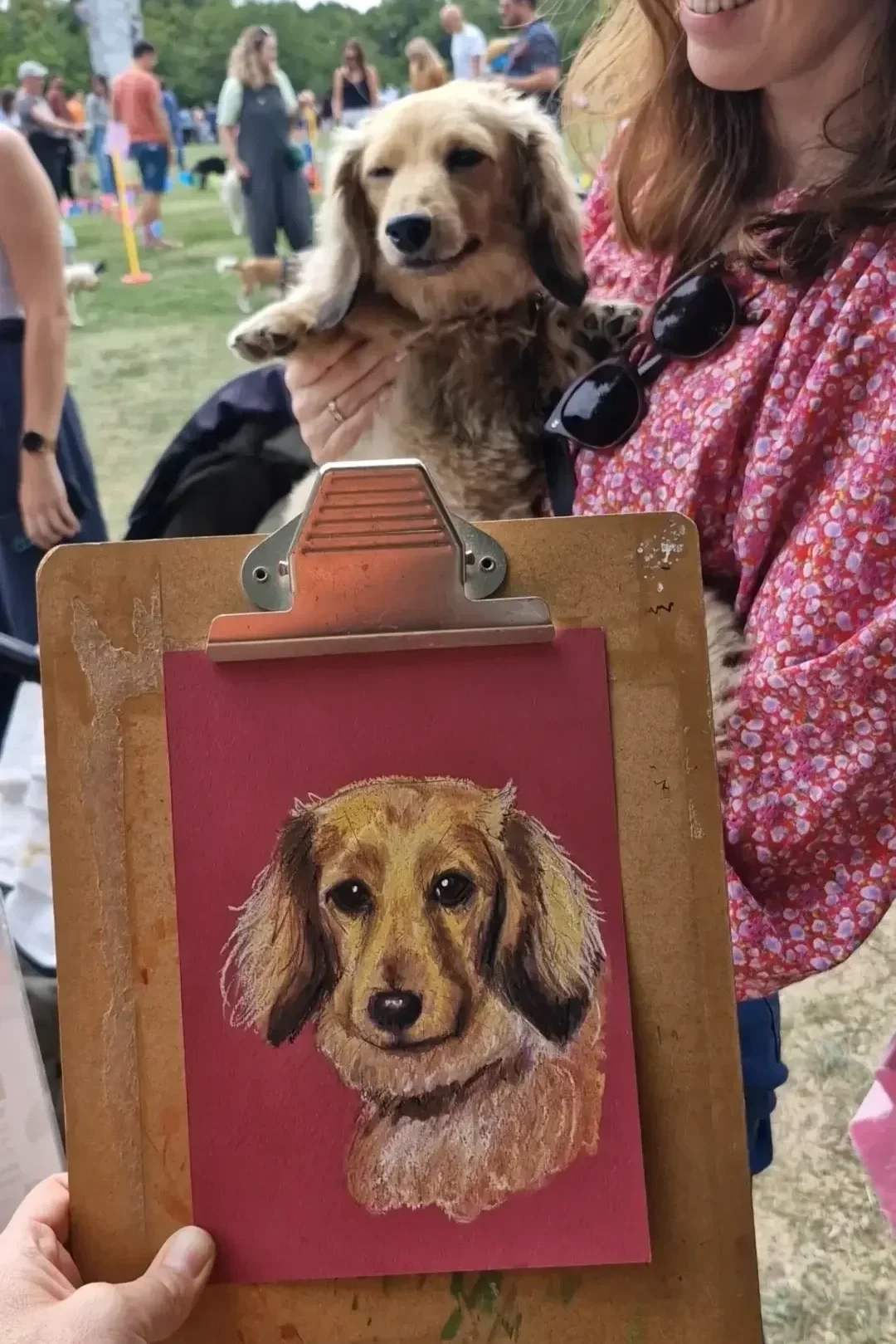 A woman holding a small dog in her arm at an outdoor event, with people in the background. In front, there's an easel with a painting of a dog that matches the dog being held. Fruity the Dachshund, Quick&Quirky A5 sketch.
