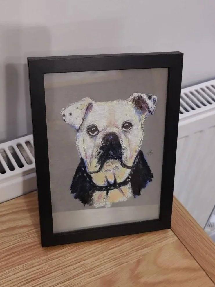 Framed chalk portrait of a tan and black bulldog with a black collar, displayed on a wooden surface.