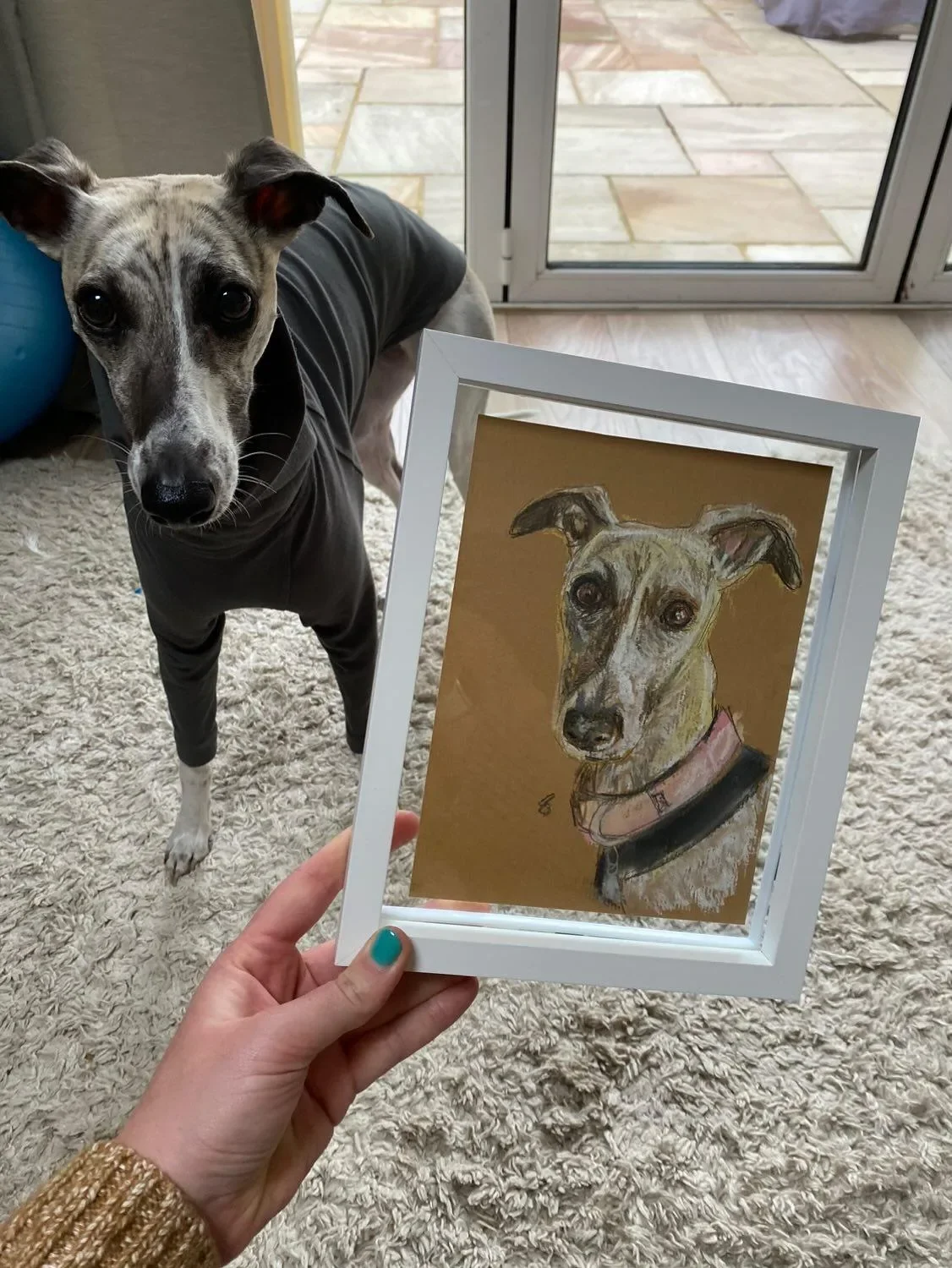 A person holding a framed chalk sketch of a dog, with the real dog looking at the camera - inside a house near a sliding glass door.