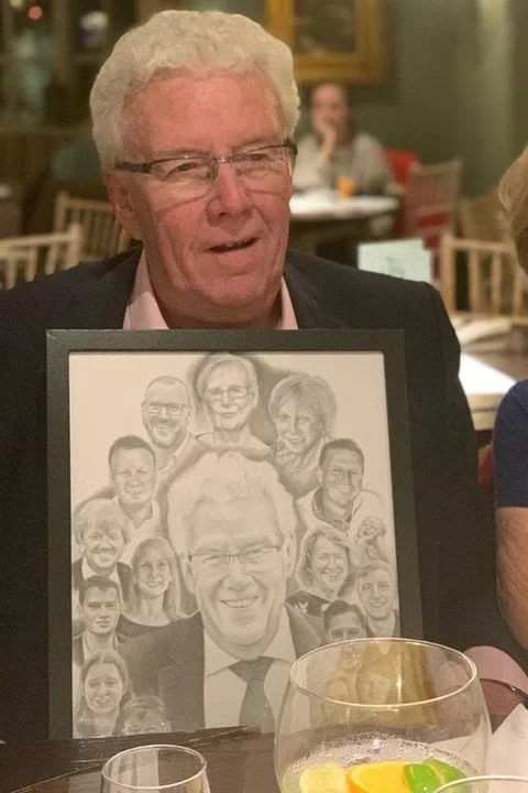 An elderly man with glasses and white hair sitting at a table in a restaurant, holding a framed collage of black and white graphite portraits - featuring himself surrounded by his family.