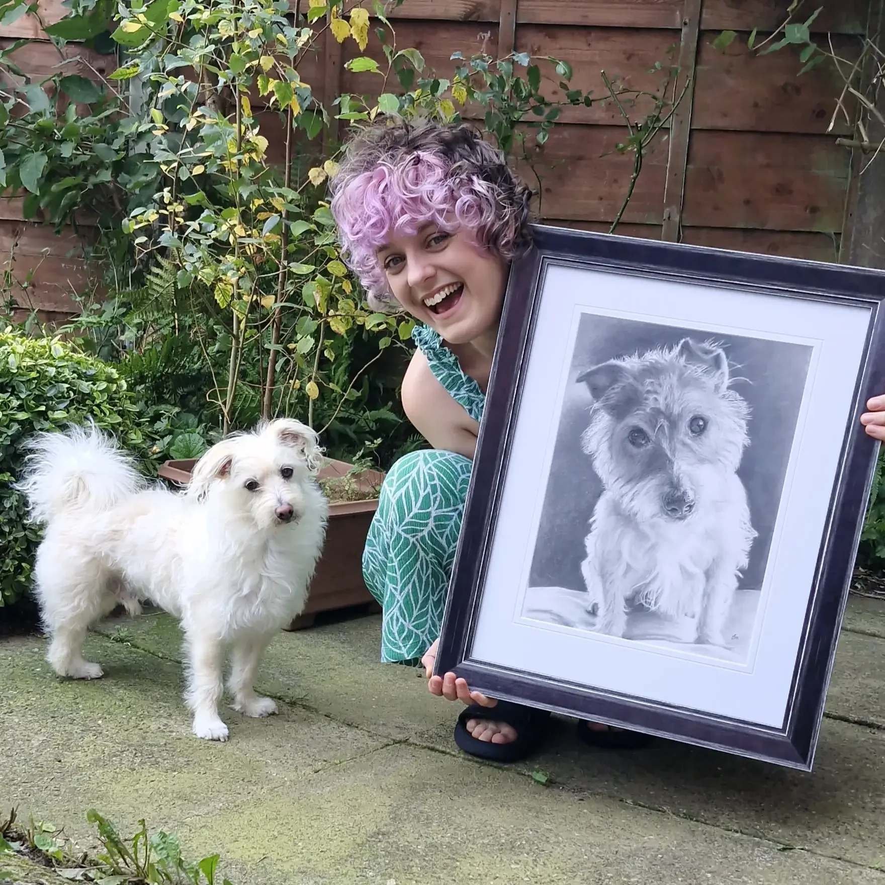 A woman with purple and black curly hair, wearing a patterned teal dress, smiling and holding a framed black-and-white portrait of a dog. She is outdoors in a garden with a small fluffy white dog standing beside her.