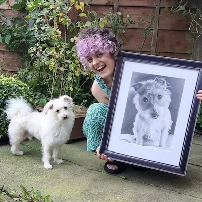 A woman with curly purple hair kneeling on a garden patio, holding a large framed black and white pencil portrait of a dog. Next to her is a small white fluffy dog standing on the ground.
