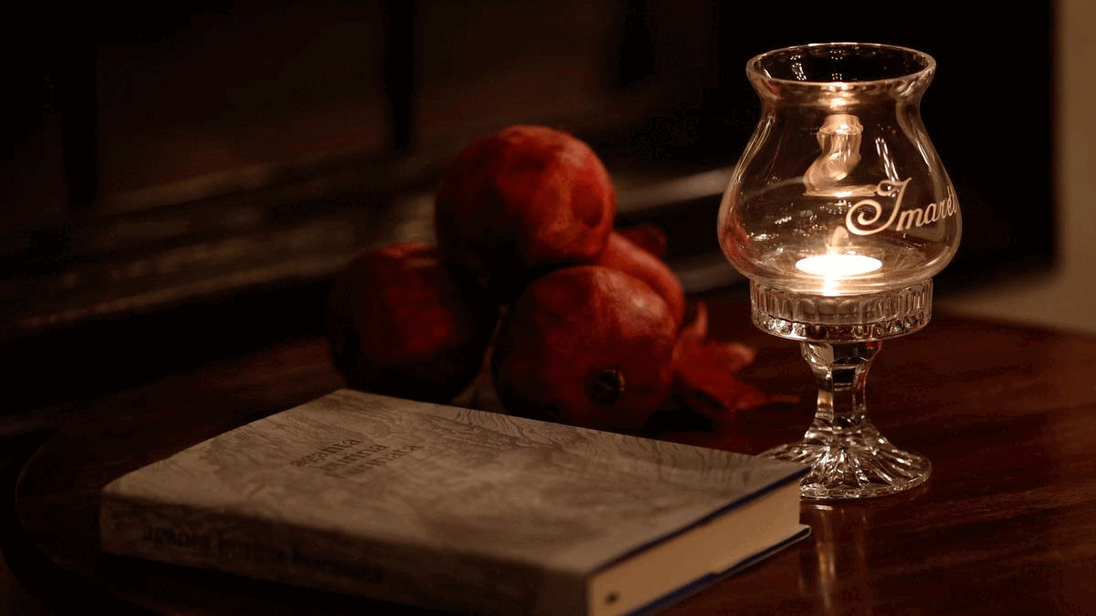 A book, a cluster of pomegranates, and a glass candle holder with a lit candle on a wooden surface.