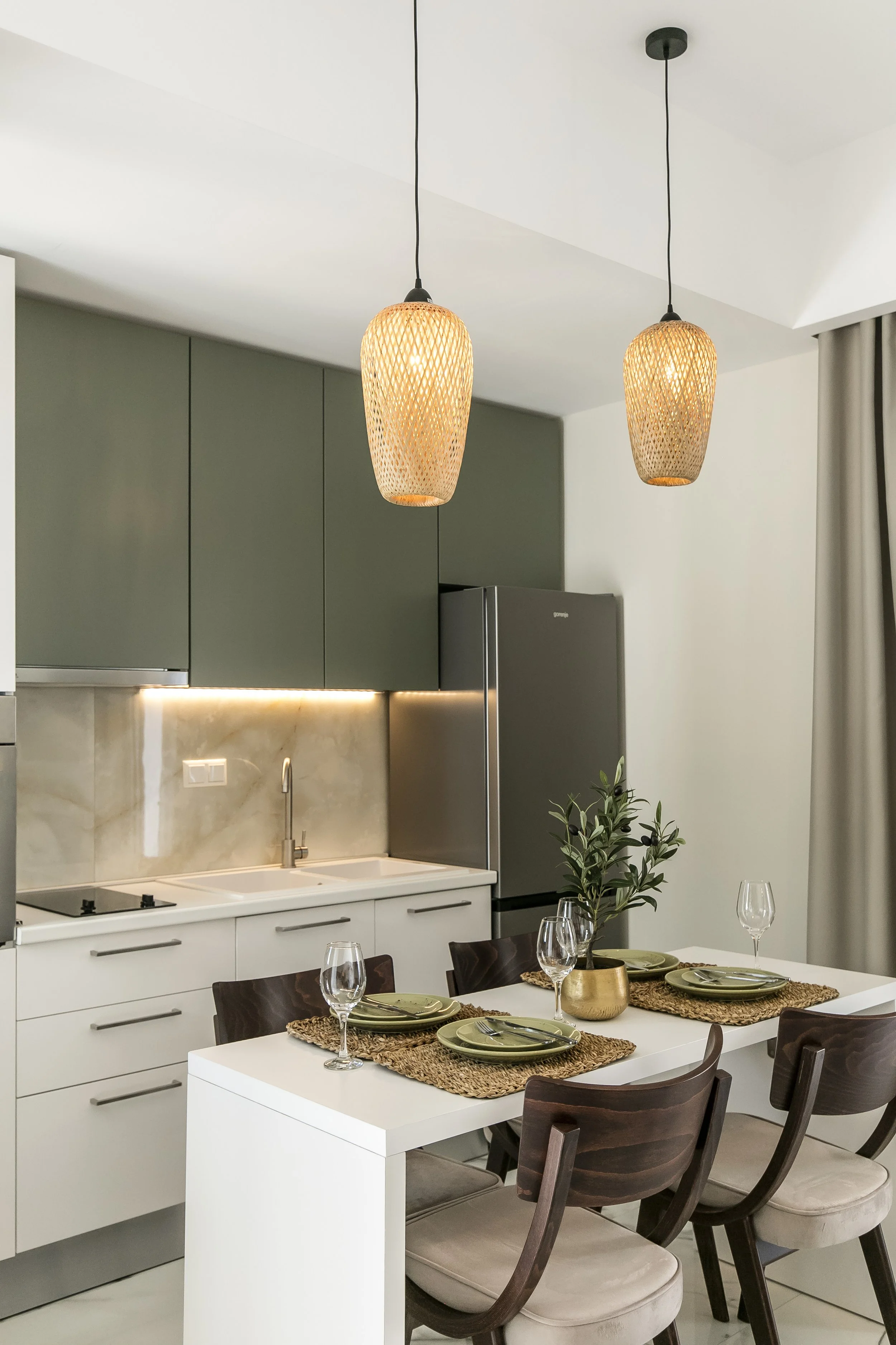 Modern kitchen and dining area with green upper cabinets, white lower cabinets, a white countertop, and a small dining table set for four with place settings, glassware, and a potted plant, lit by hanging wicker pendant lights.