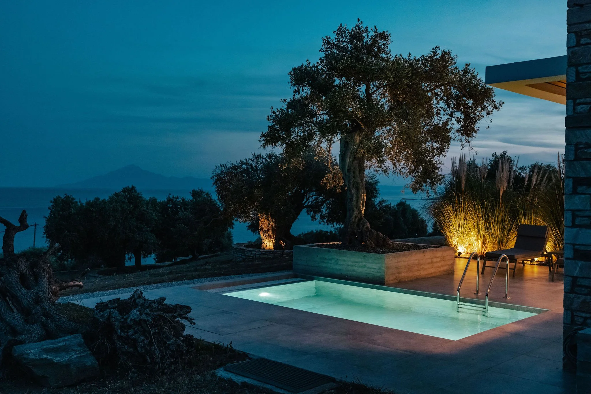 Outdoor swimming pool illuminated at night, surrounded by trees, with lounge chairs and decorative lighting, overlooking a distant mountain range.