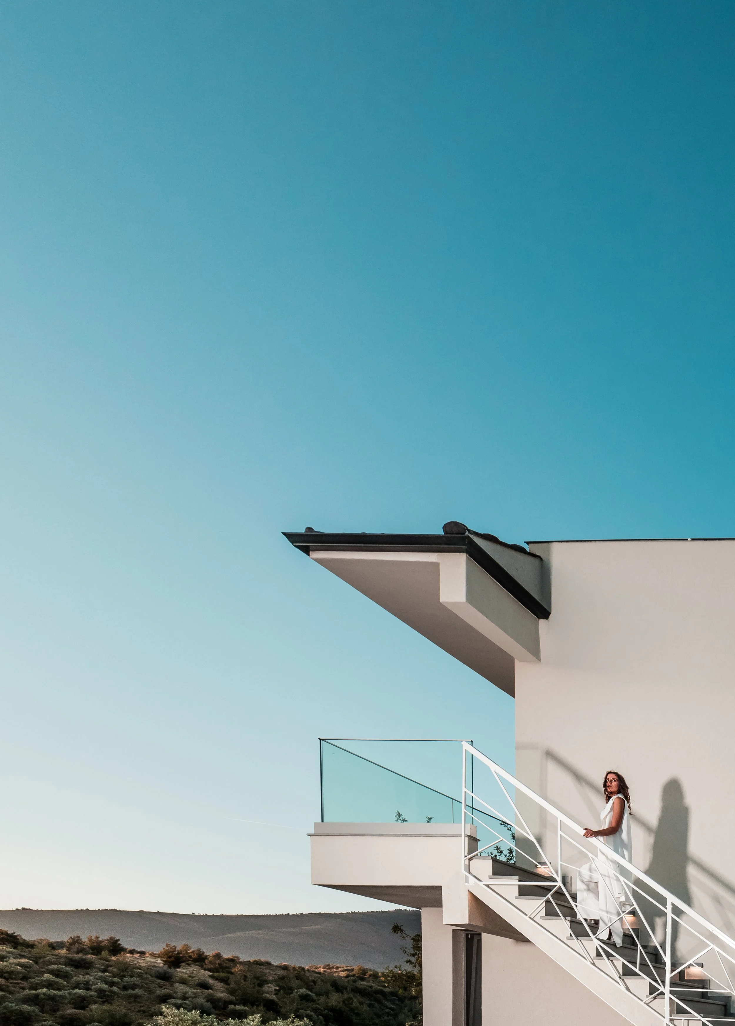 A woman in white dress standing on exterior staircase of a modern white building with glass balcony, overlooking a landscape with hills under a clear blue sky.