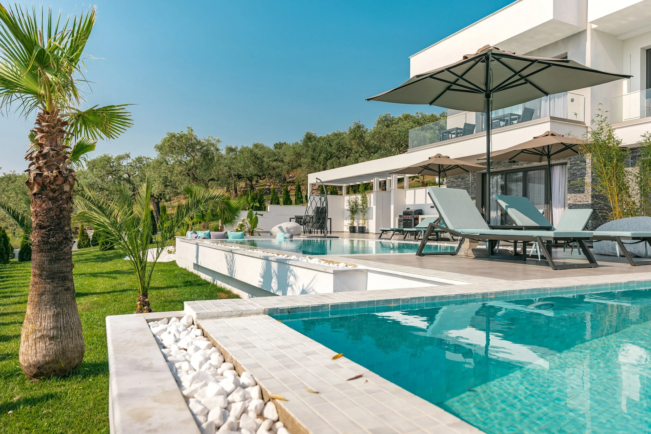 Modern outdoor swimming pool area with lounge chairs, umbrellas, and green landscaped surroundings with trees and grass.