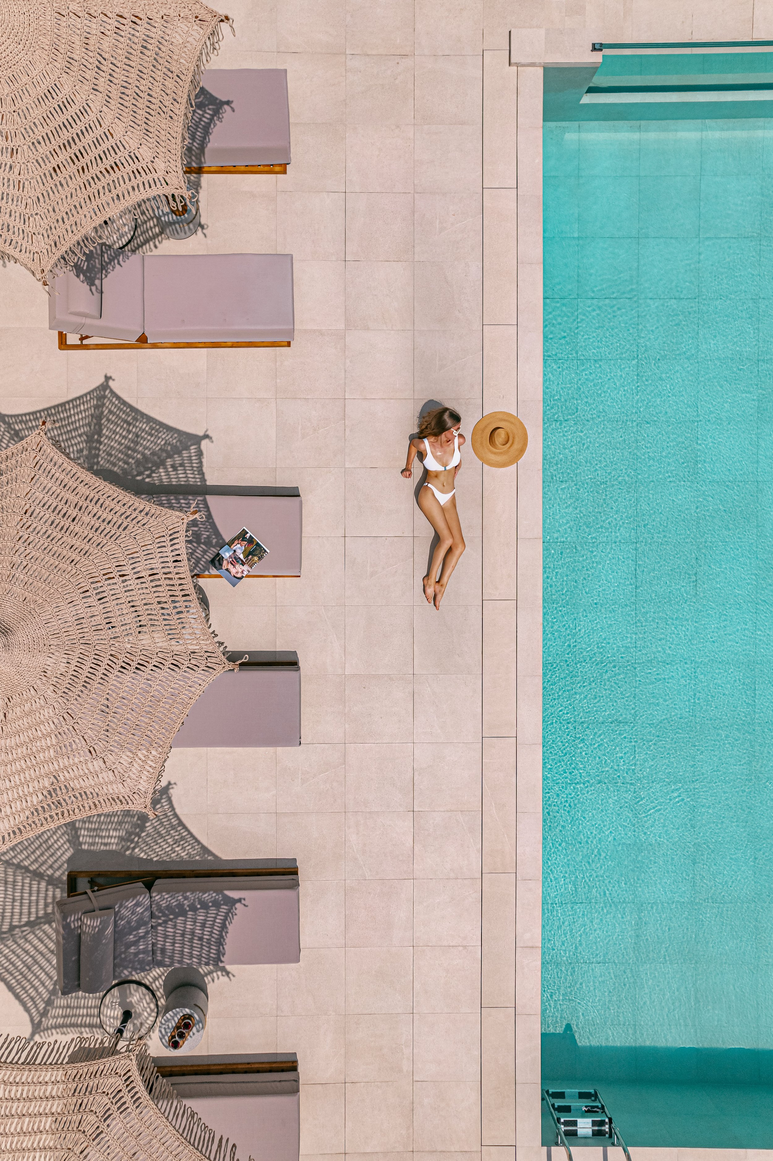 A woman in a white bikini lies on a poolside lounge chair near the pool, with a wide-brimmed straw hat beside her, on a tiled pool deck. Two umbrellas provide shade over two other lounge chairs, with a magazine resting on one of them.