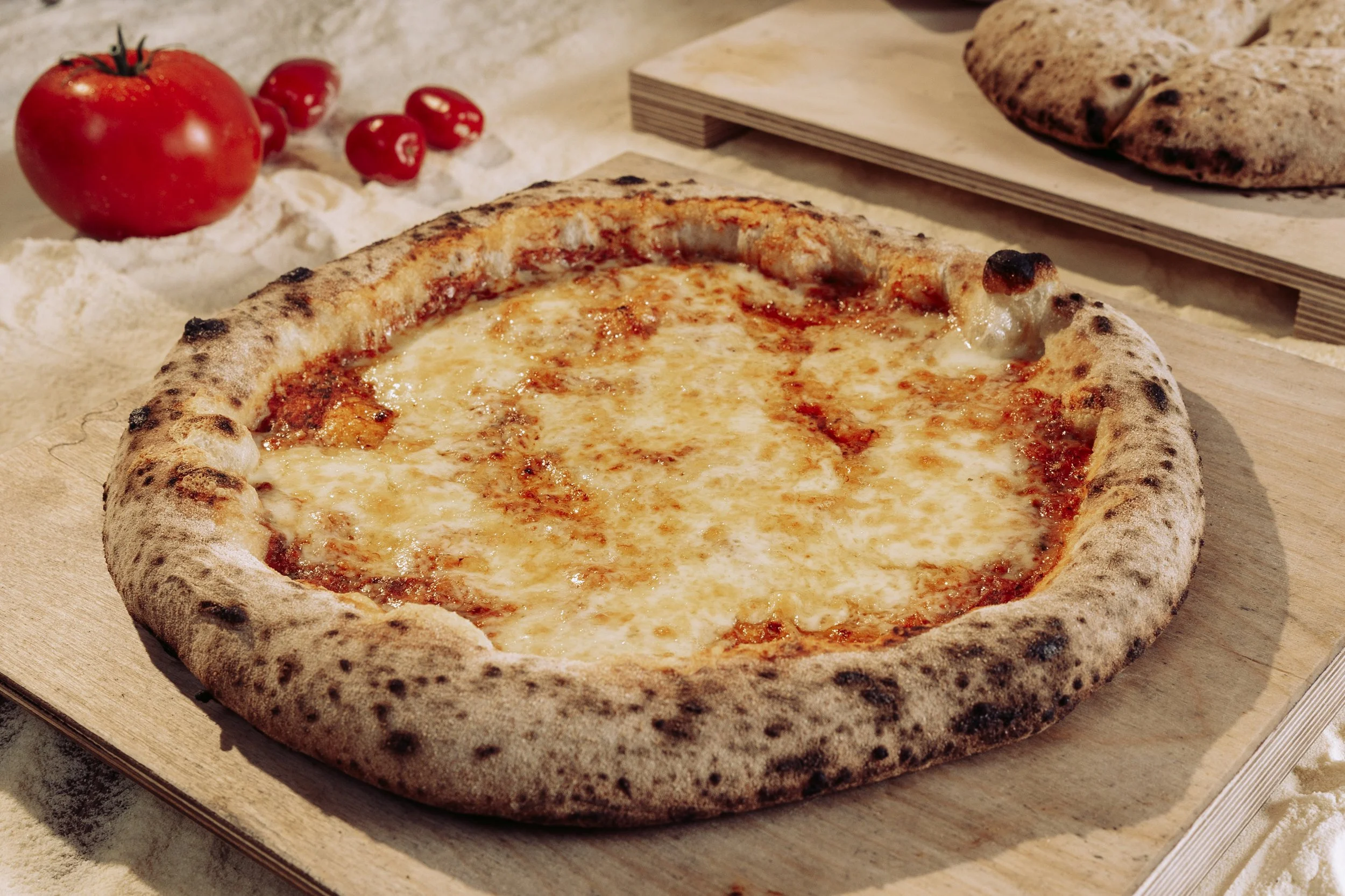 A cheese pizza with a golden crust on a wooden pizza board, with tomatoes and bread in the background.