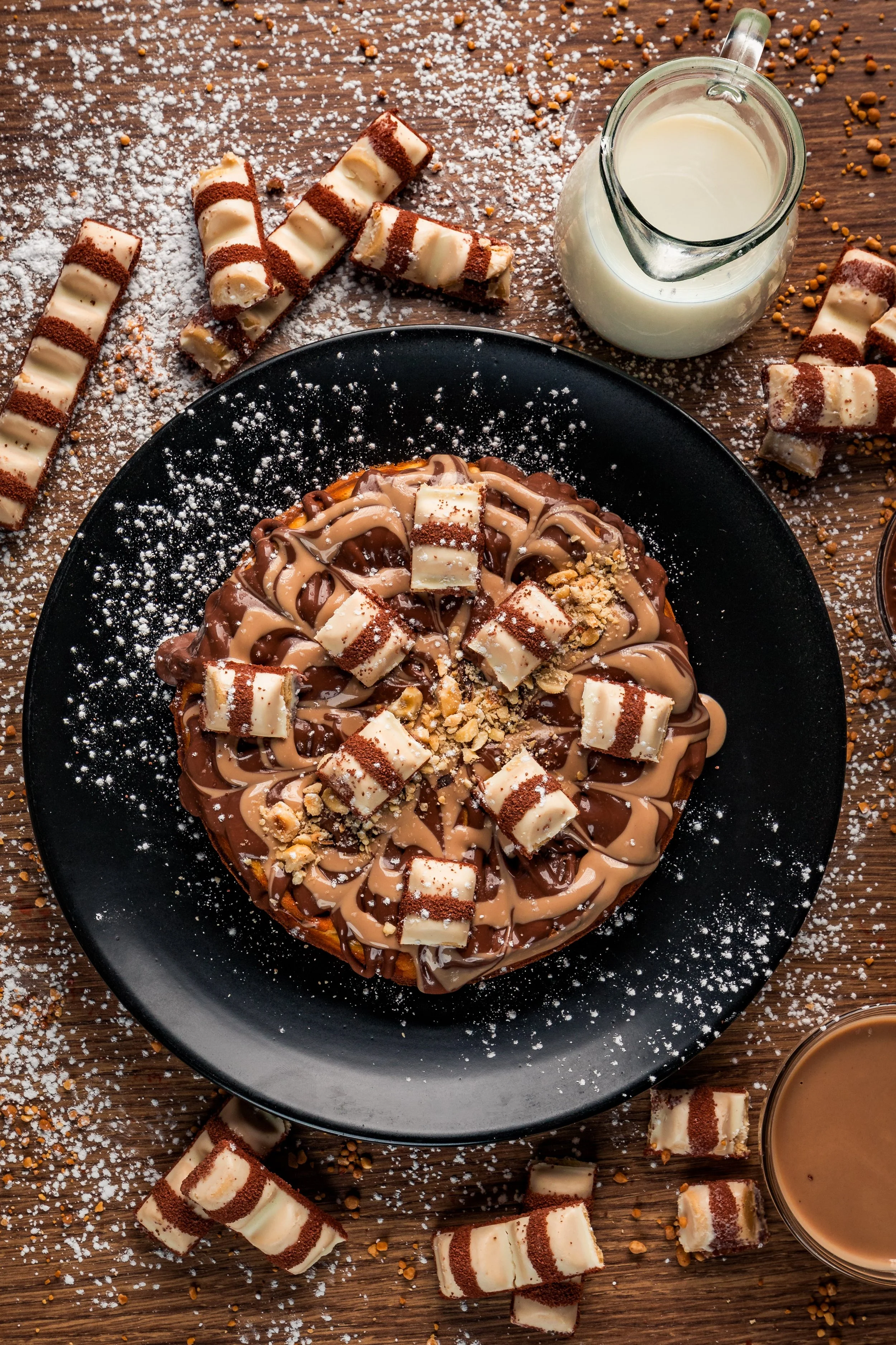 Chocolate and white chocolate dessert topped with white chocolate cubes and crushed nuts, placed on a black plate, surrounded by rolled chocolate wafer cookies, a glass jug of milk, and cups of coffee on a wooden table with powdered sugar and crushed