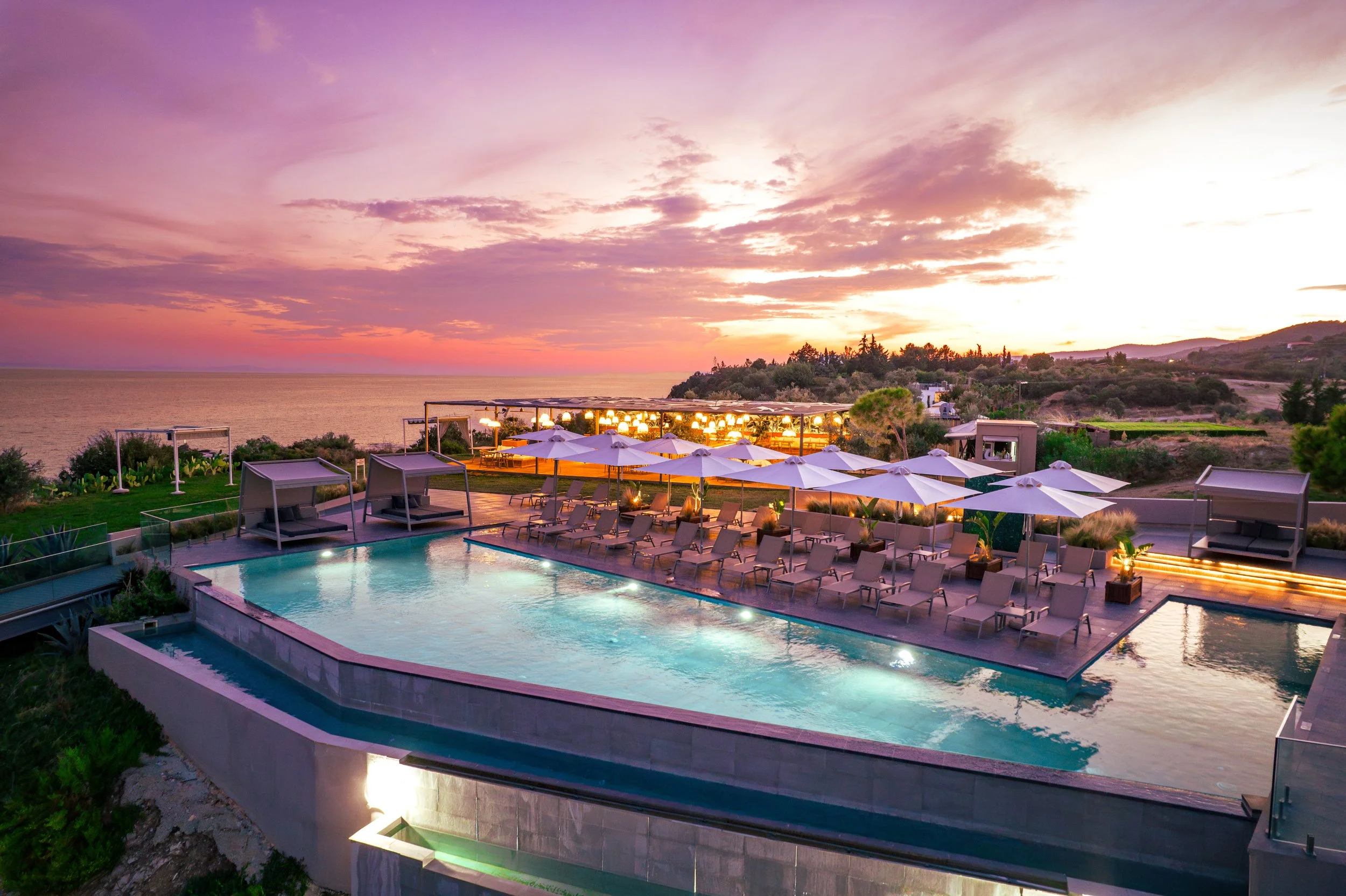 A luxurious seaside poolside area at sunset with a large infinity pool, lounge chairs with umbrellas, shaded cabanas, and an ocean view in the background under a colorful sky.