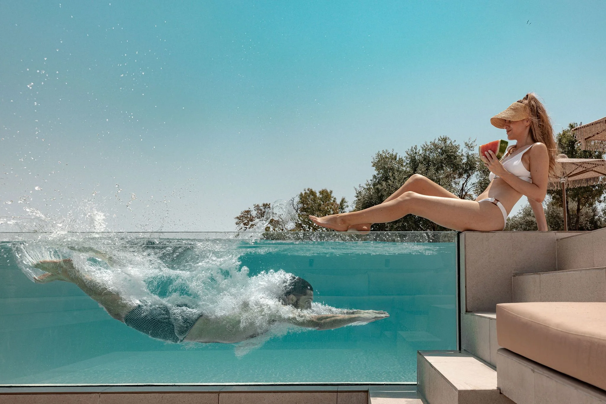 A woman in a white bikini and straw hat sitting on poolside steps, holding a slice of watermelon, while a man swims underwater in a clear pool beneath her.