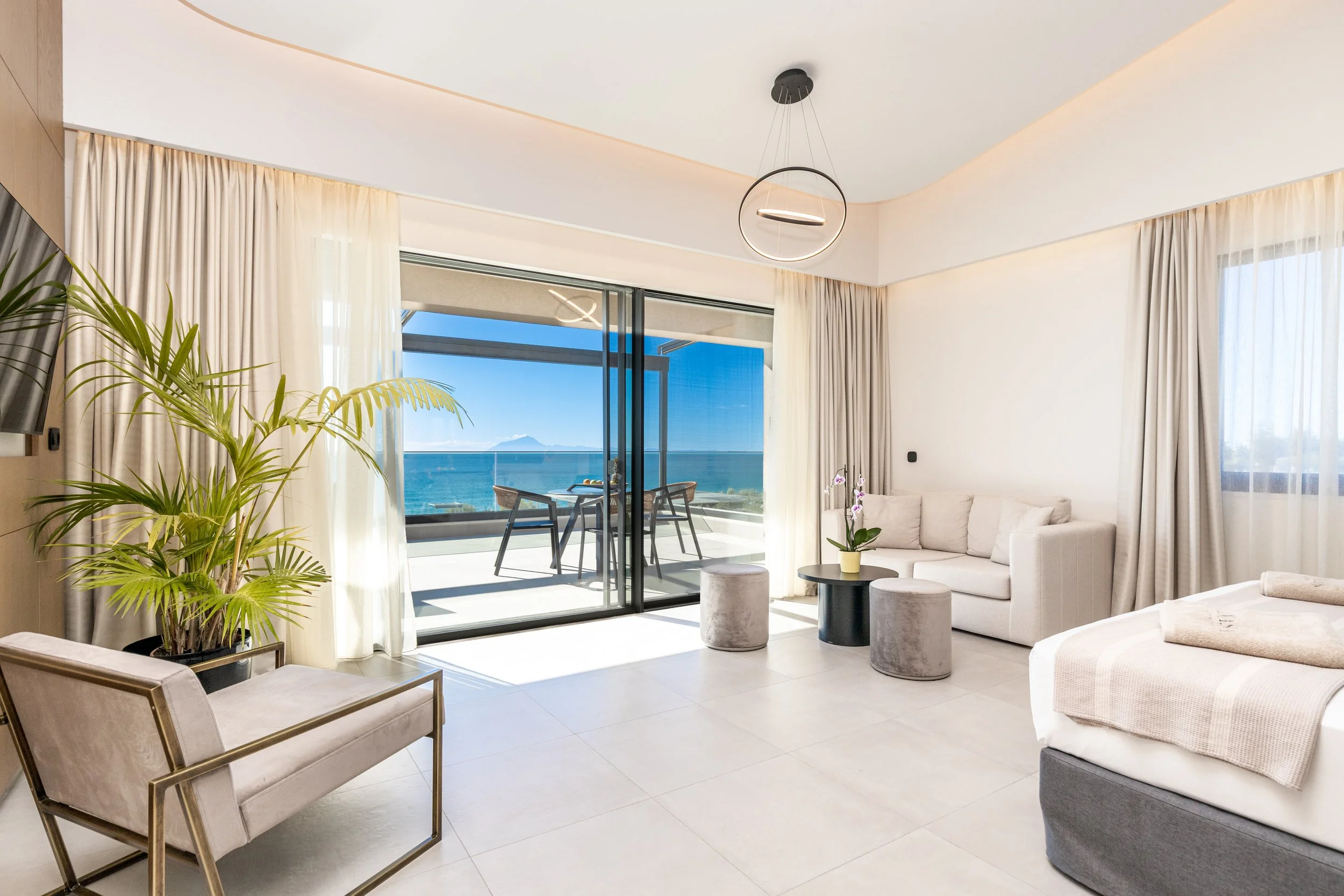Modern hotel room with white sofa, bed, potted orchids, armchair, a balcony with ocean view, and contemporary lighting.