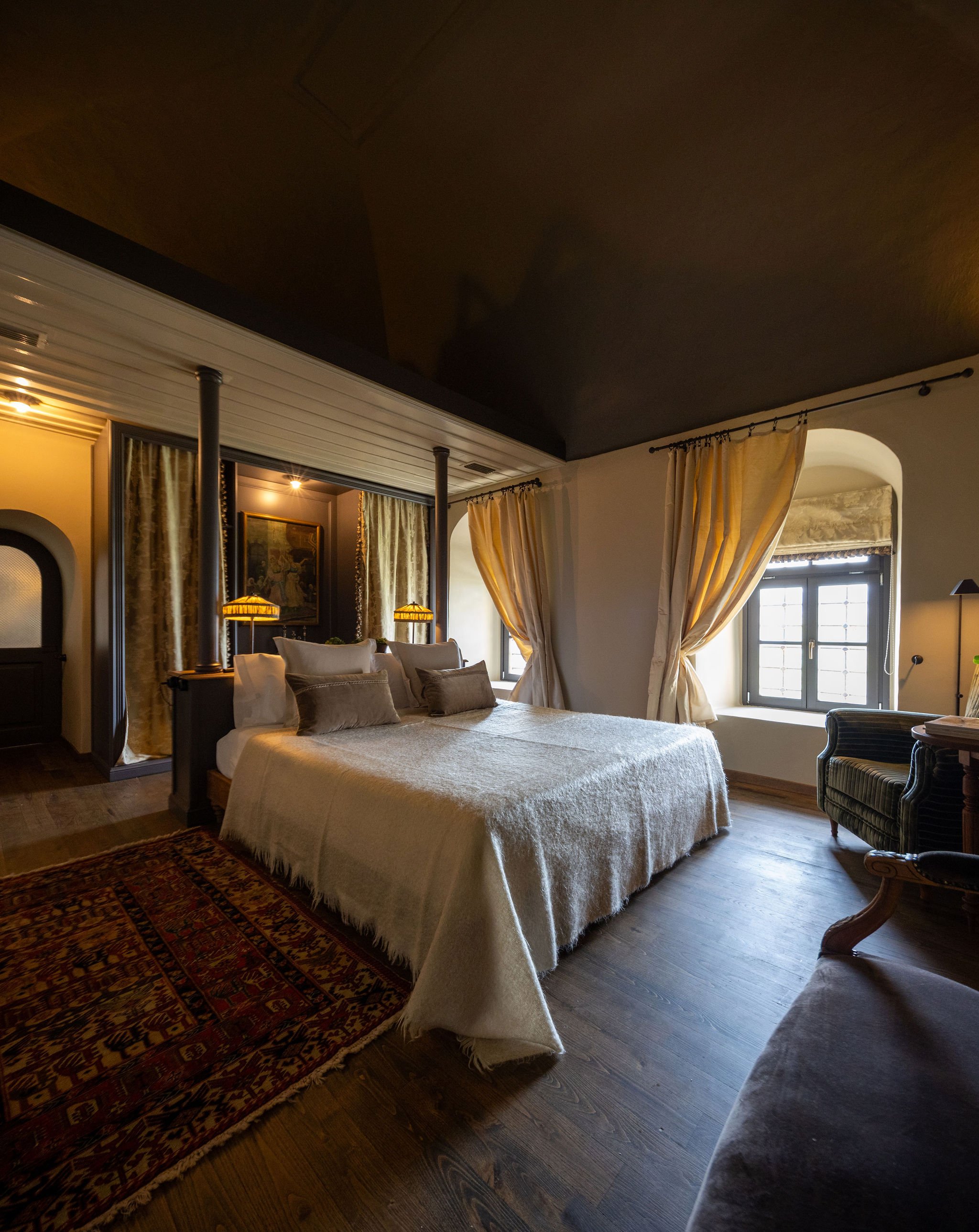 A cozy bedroom with a bed covered in a beige blanket, decorative pillows, and a wooden floor. There are windows with cream curtains, a sitting armchair, and warm lighting.