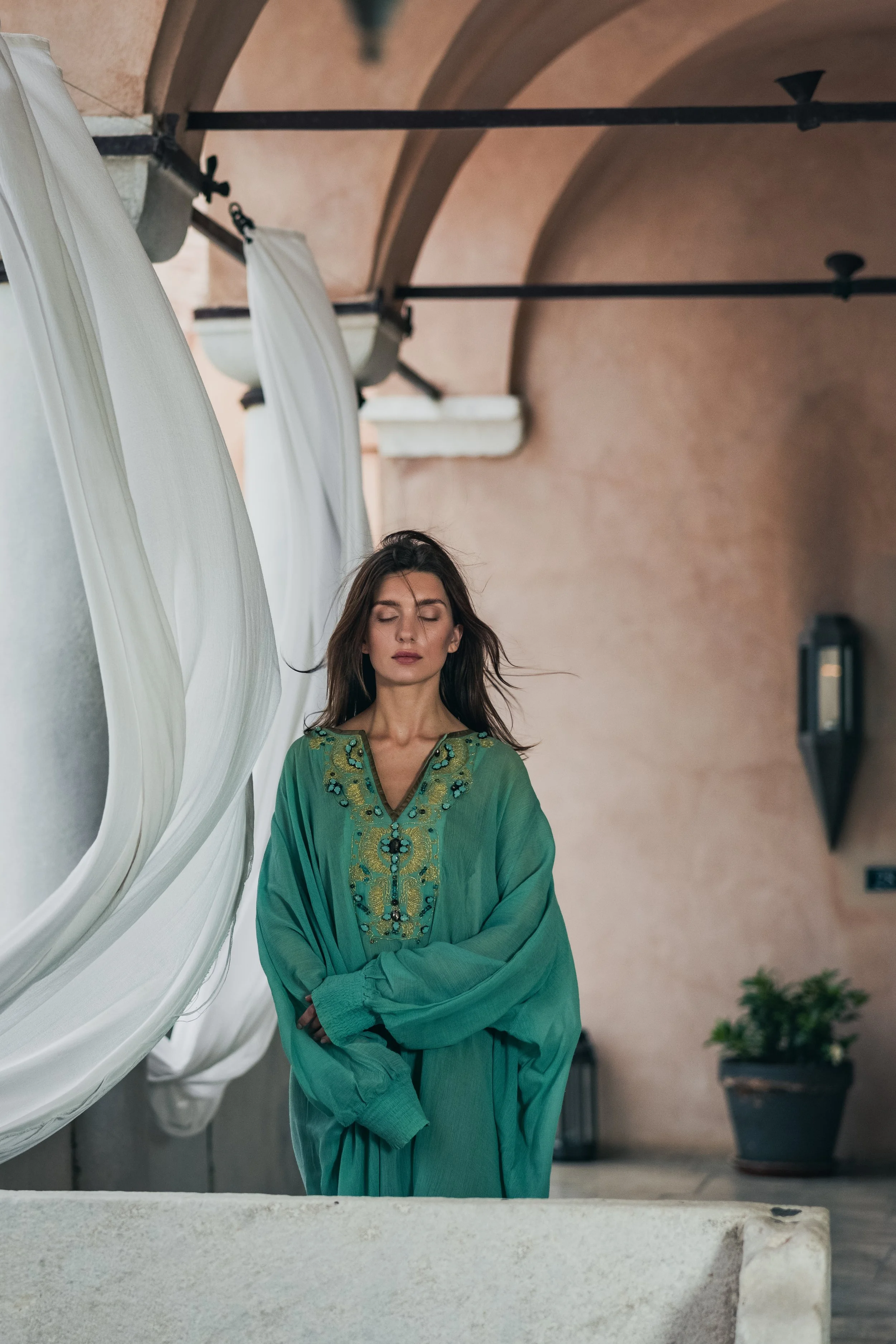 A woman with long brown hair and closed eyes stands in front of a stone wall, wearing a green embroidered tunic, with white curtains and an arched ceiling in the background.
