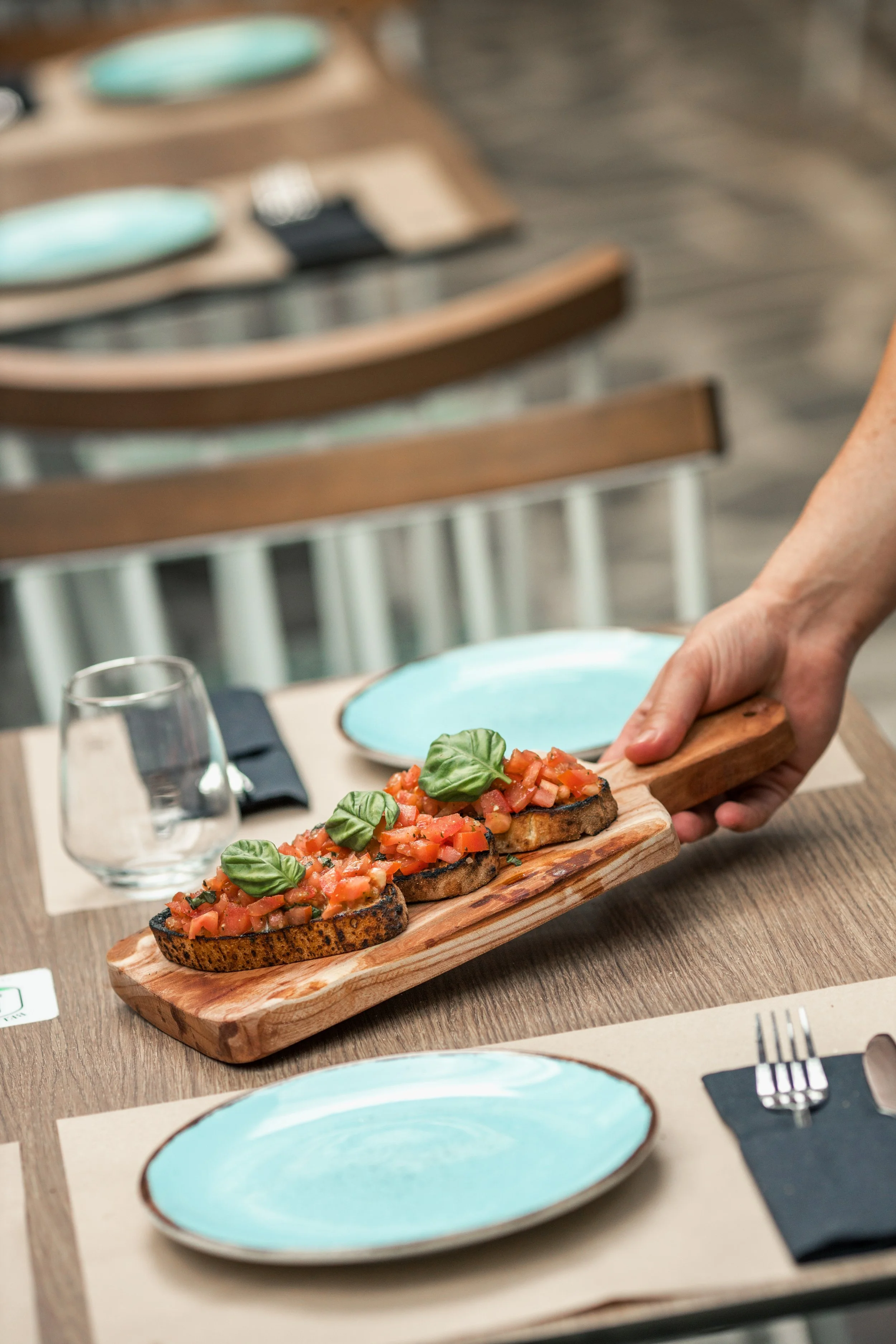 A hand holding a wooden serving board with three pieces of bruschetta topped with diced tomatoes and basil leaves, on a dining table with turquoise plates and a wine glass.
