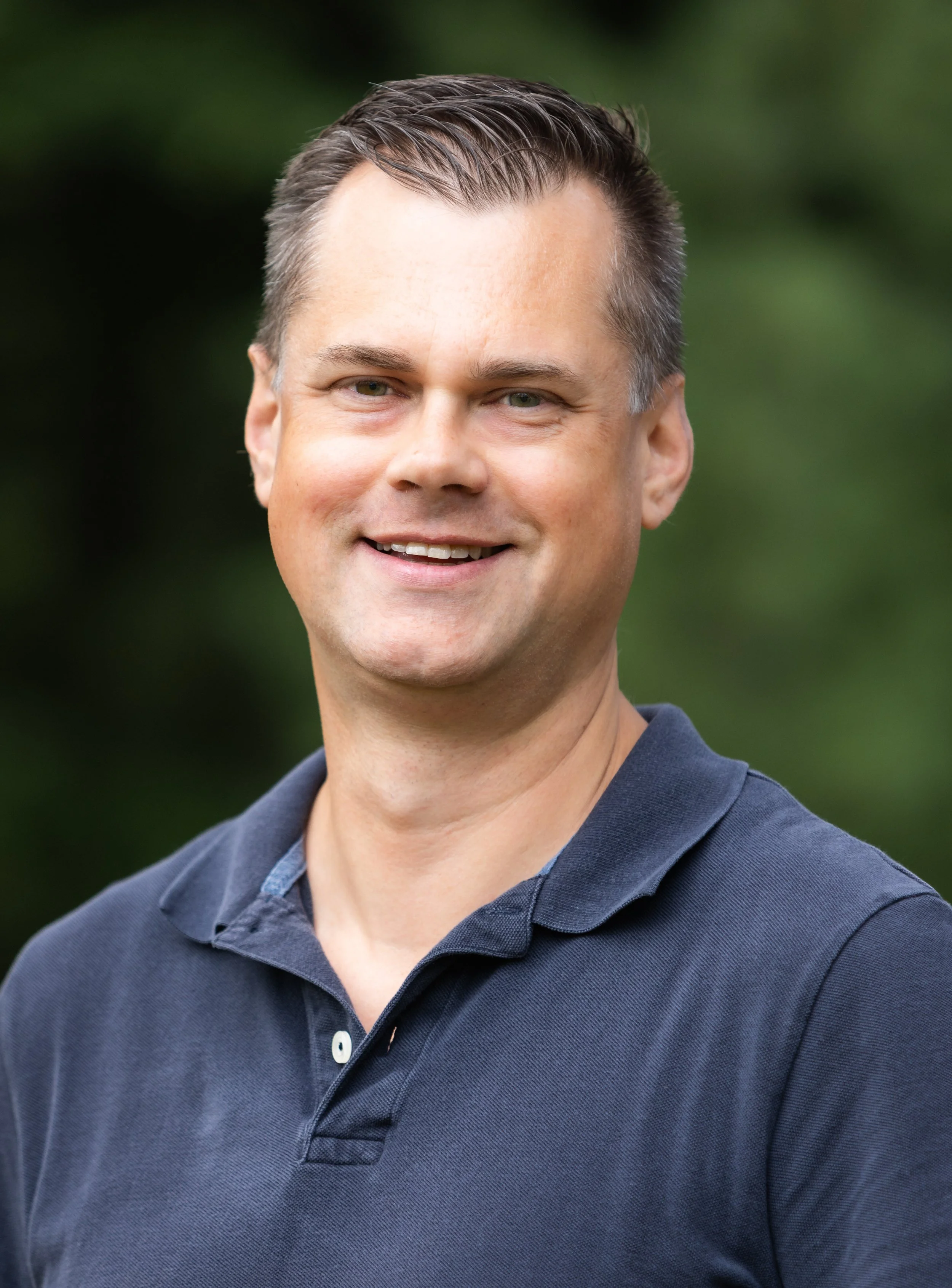 A man with short brown hair, wearing a dark polo shirt, smiling outdoors with a blurred green background.