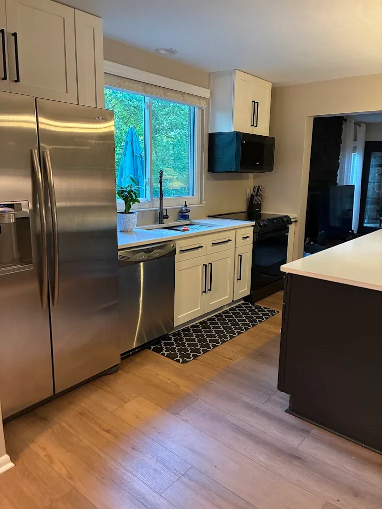 Modern kitchen with stainless steel refrigerator, dishwasher, and oven, white cabinets, a window with a view of trees, and a black and white patterned rug on wood flooring.