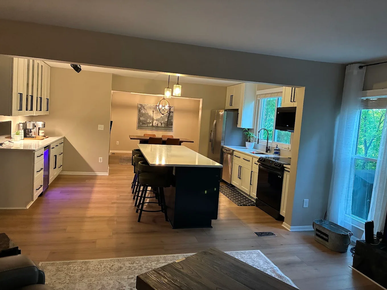 Modern kitchen with island, white cabinets, black appliances, large window, and sliding glass door leading outside, hardwood floors, and dining area in the background.