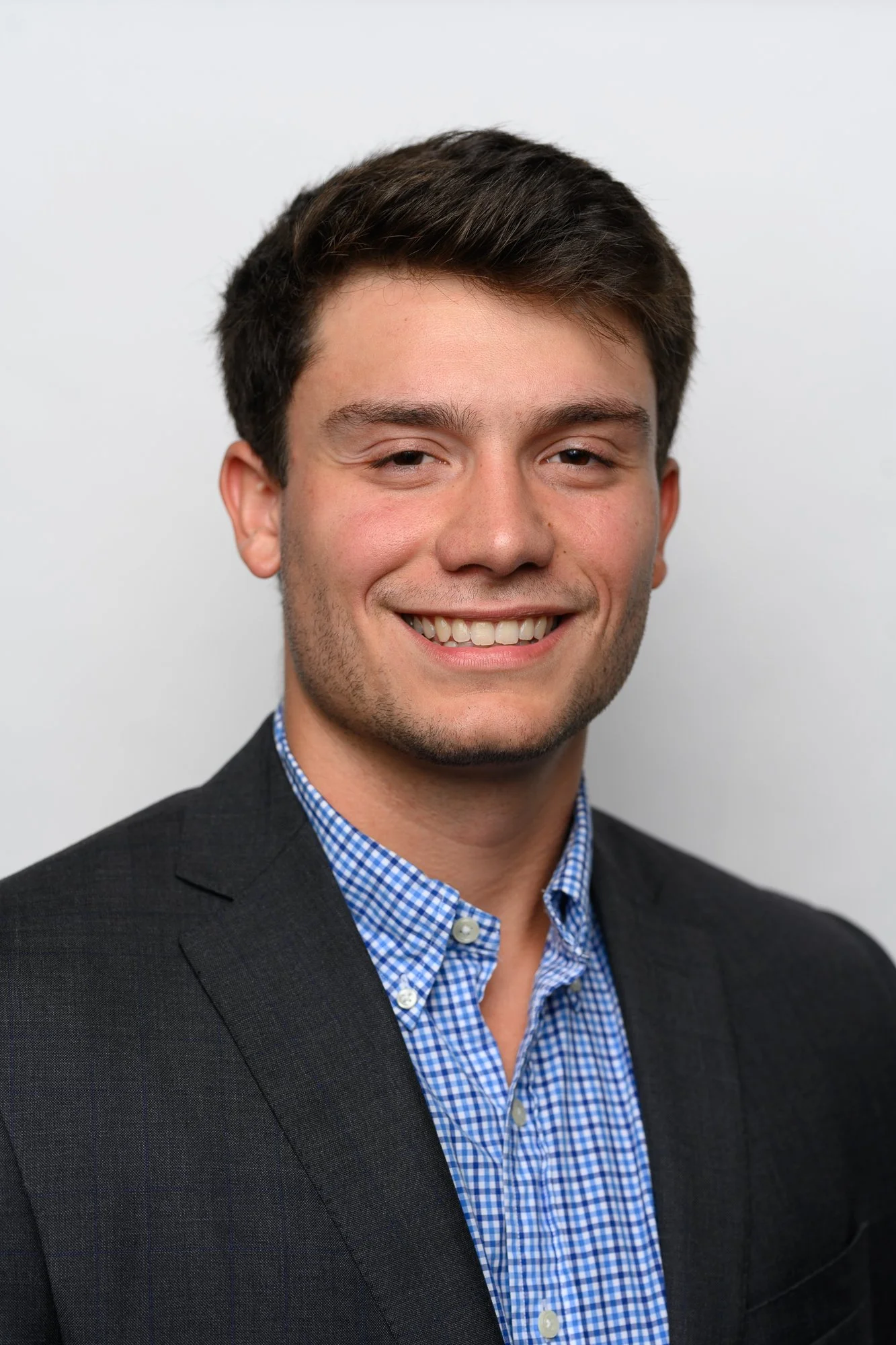 A young man with dark brown hair, smiling, wearing a dark suit jacket and a blue checkered shirt, against a plain white background.