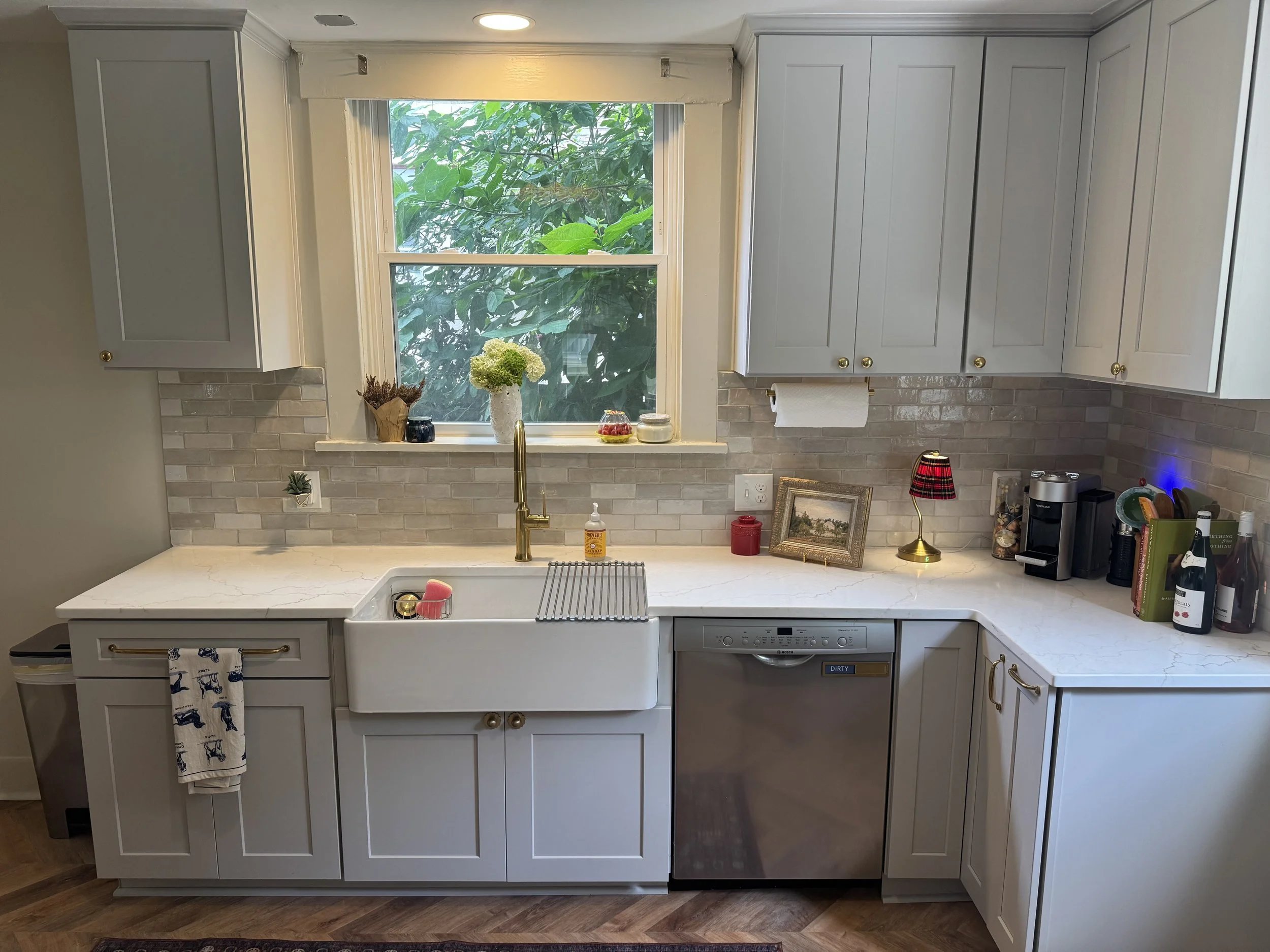 A modern kitchen with white cabinets, a large window with greenery outside, a kitchen sink, and various decorative items on the countertop.