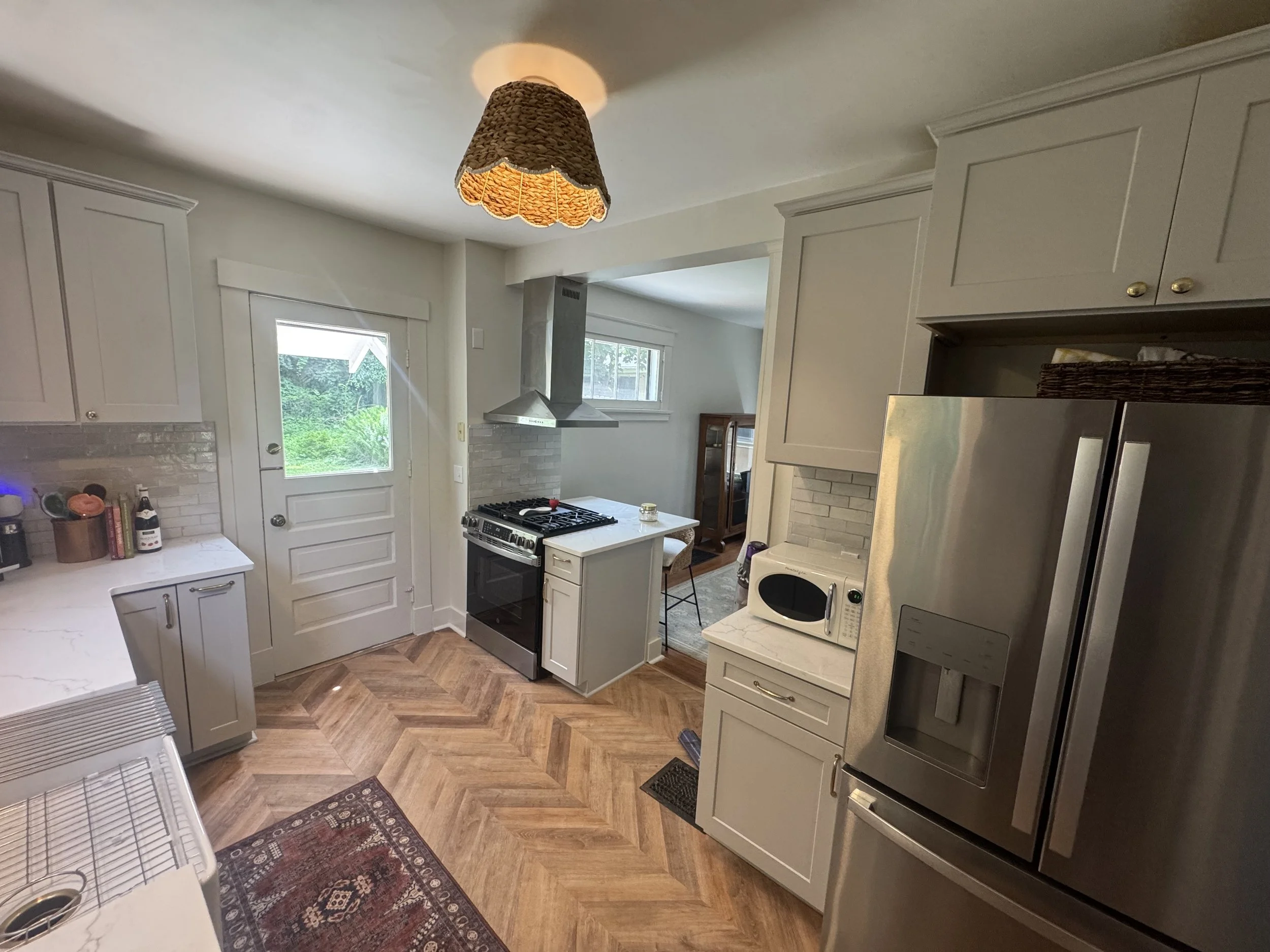 A kitchen with white cabinets, a stove, a microwave oven, stainless steel refrigerator, and wooden flooring. There are windows showing greenery outside, and a wicker lampshade hanging from the ceiling.
