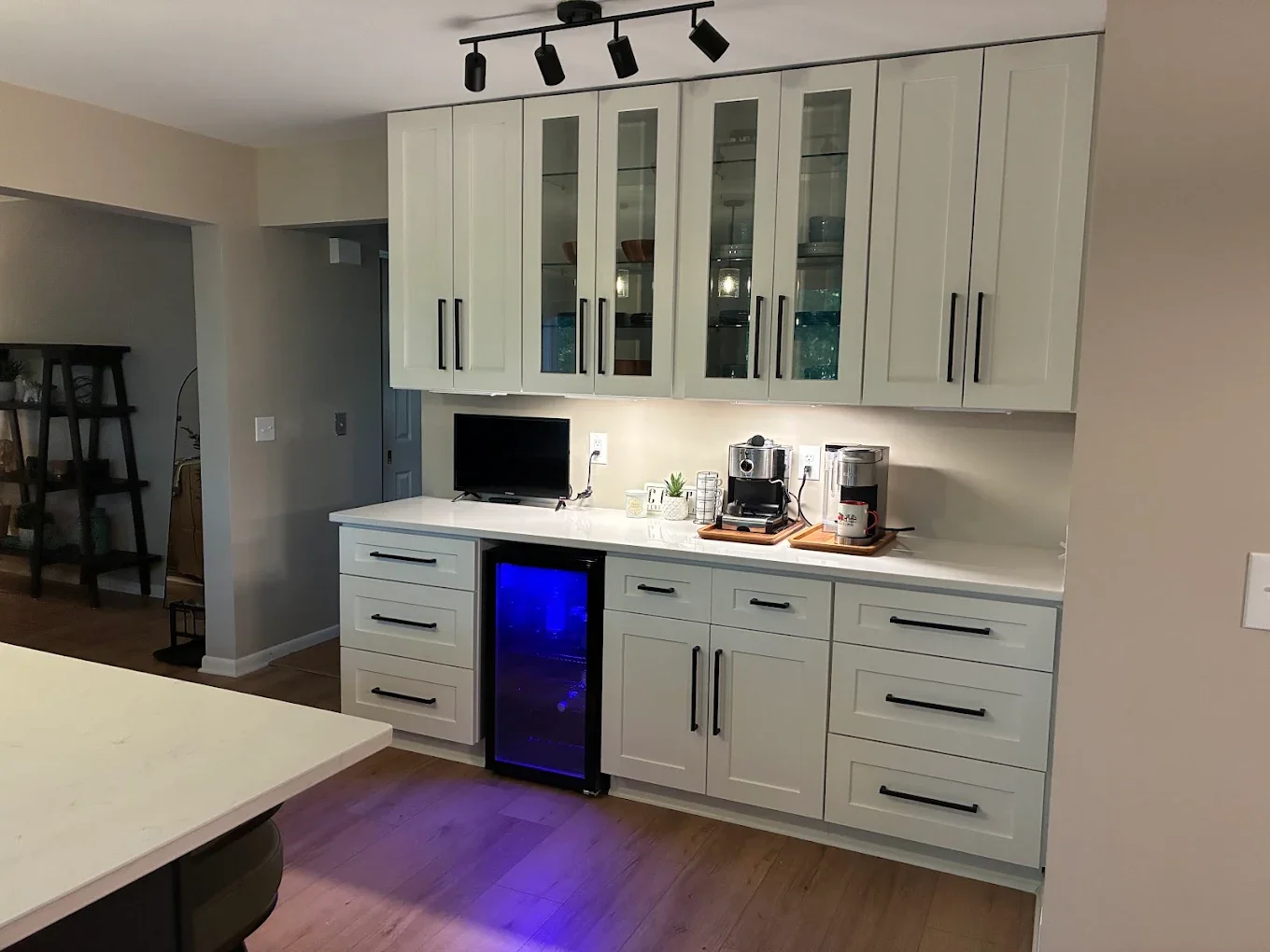 Kitchen with white cabinets, black handles, a small TV, coffee machine, espresso machine, mini wine cooler with blue light, and coffee mugs on the counter.