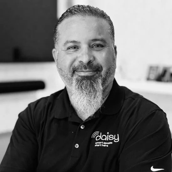 A man with a beard and mustache smiling, wearing a black polo shirt with a company logo and slogan, sitting indoors.