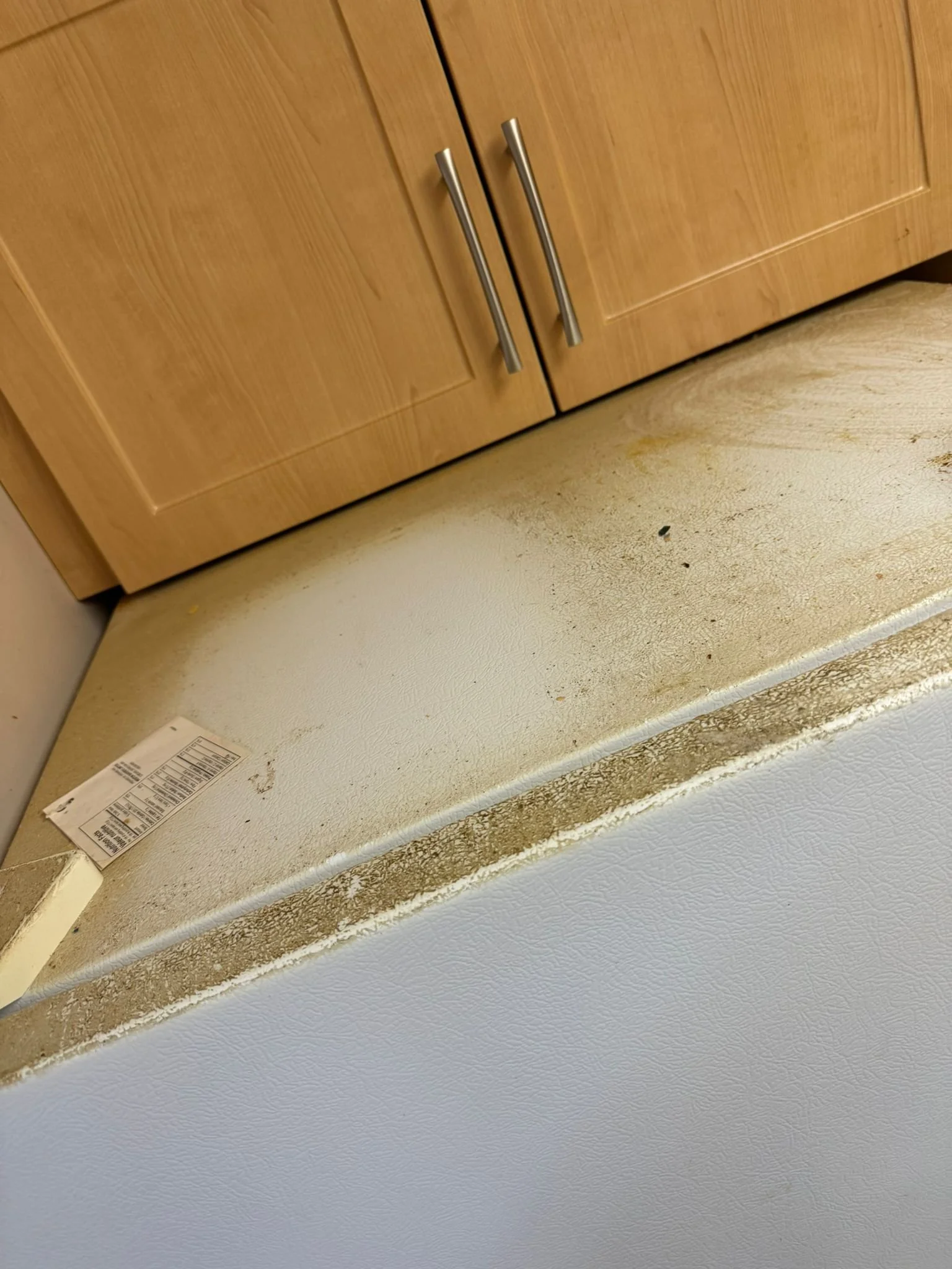 Dirty kitchen counter with a sticker, above a white textured refrigerator, with wooden cabinets in the background.