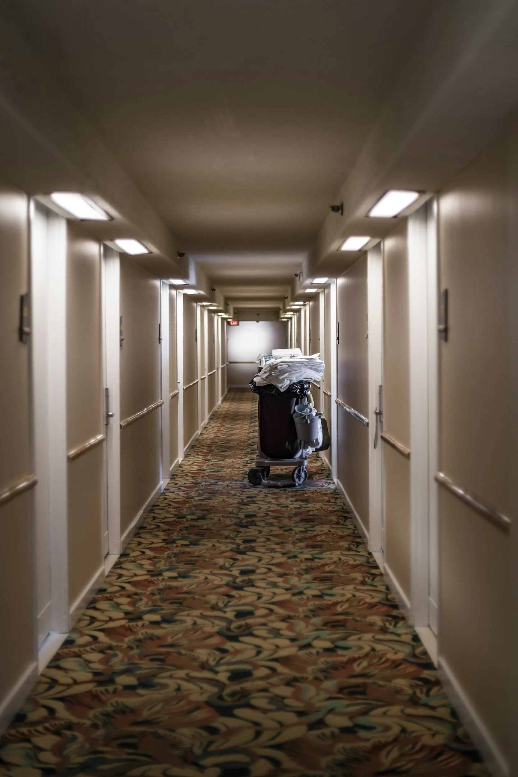 Hotel hallway with patterned carpet and multiple closed doors, with a cleaning cart piled with white linens in the middle.