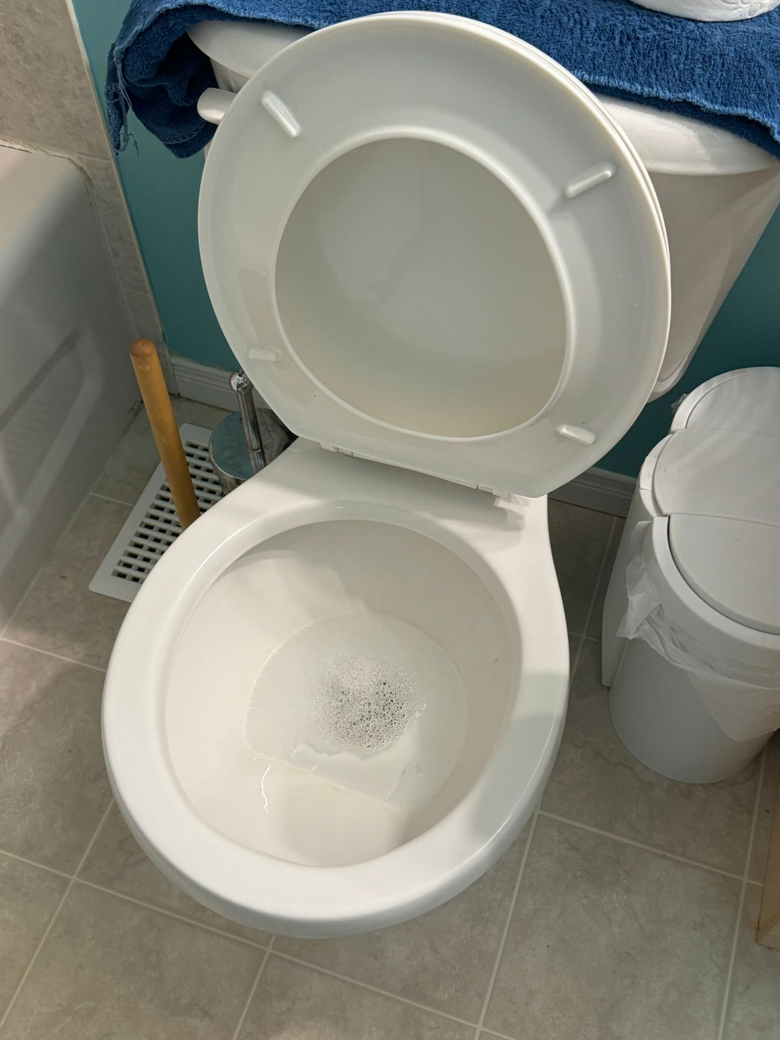 An open toilet with water inside, a blue towel draped over a fixture, a plunger, a vent, and a trash can with a trash bag in a bathroom with tiled floor.