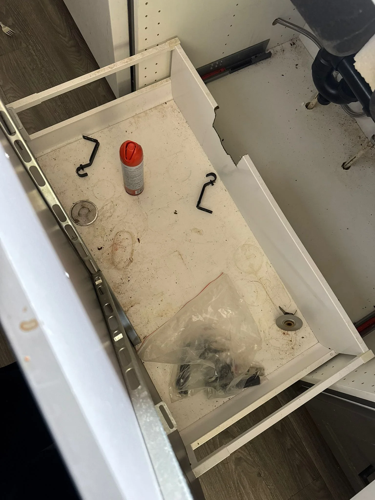 Inside a partially disassembled washing machine drawer, with dirty surfaces, some small tools, a can of lubricant, and a plastic bag containing a black component.