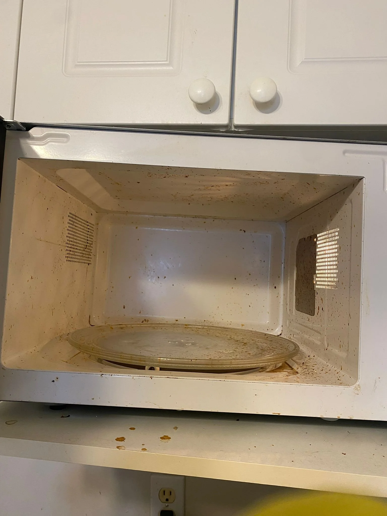 Empty microwave oven with food splatters inside, above a countertop with a power outlet.