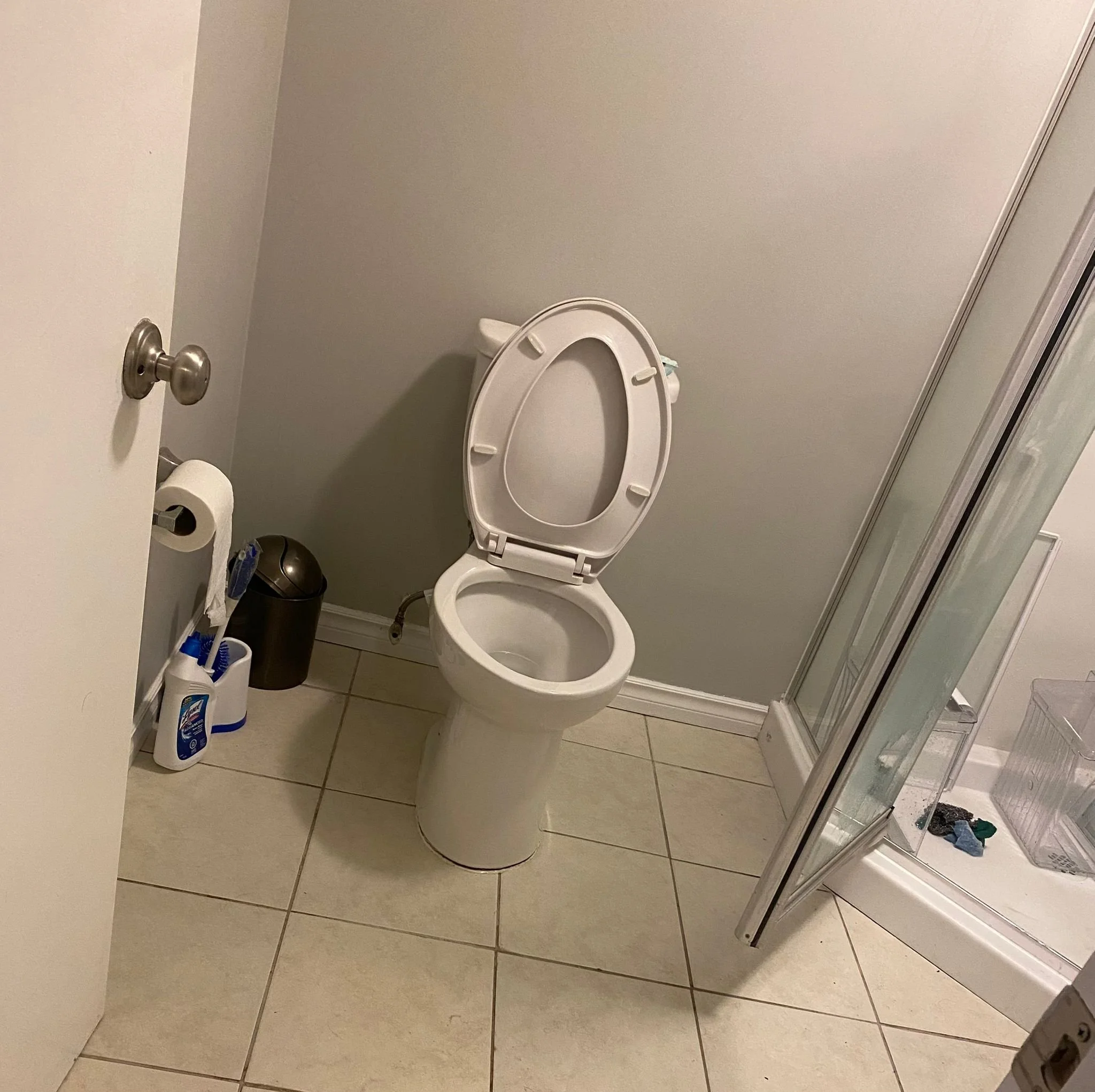 A bathroom with a toilet, a small trash bin, a toilet brush, toilet paper, a shower with glass door, and a box of cleaning supplies on the floor.