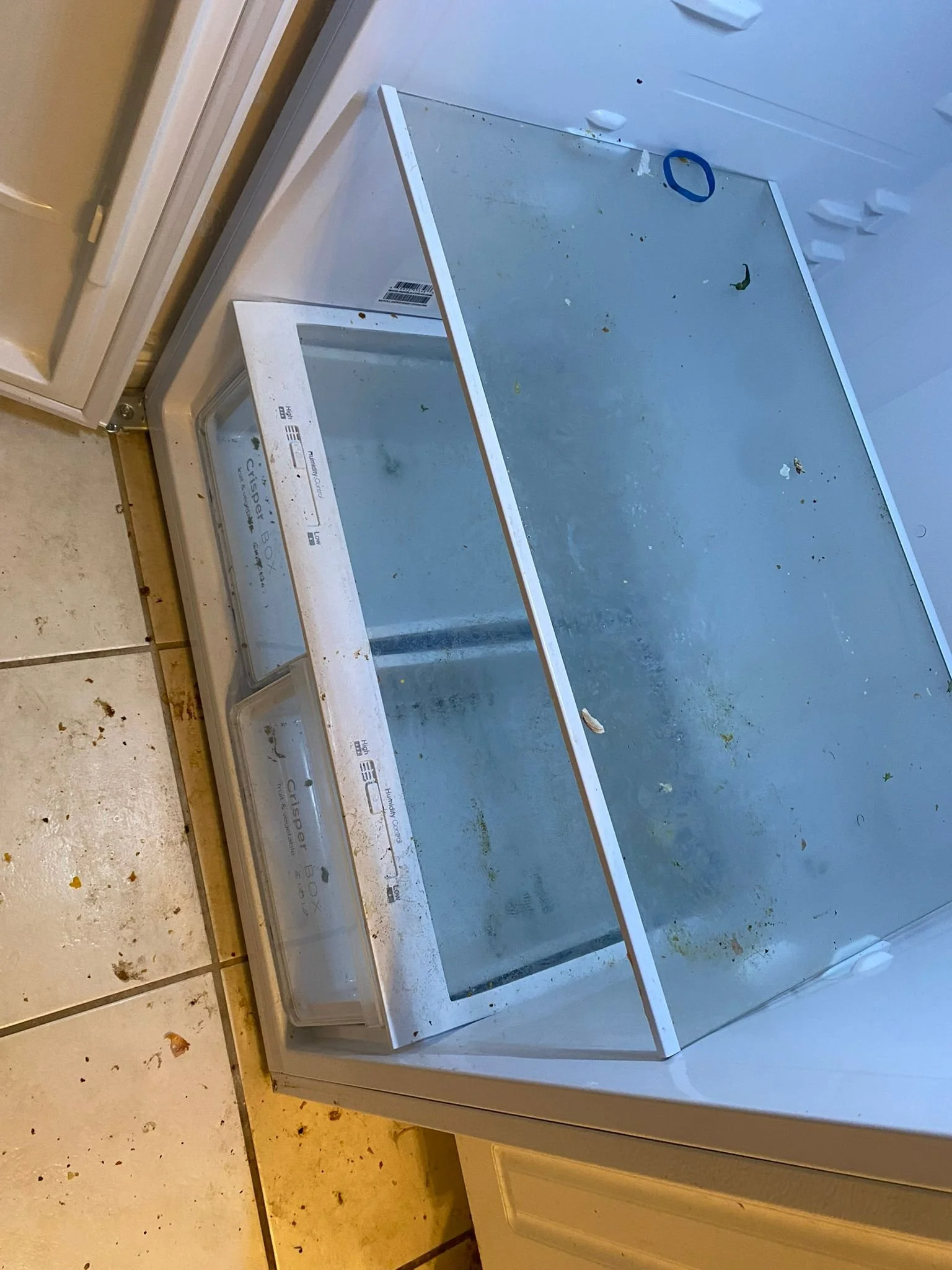Open, empty freezer with dirt and small debris inside, in a kitchen with tiled floor and white cabinets.