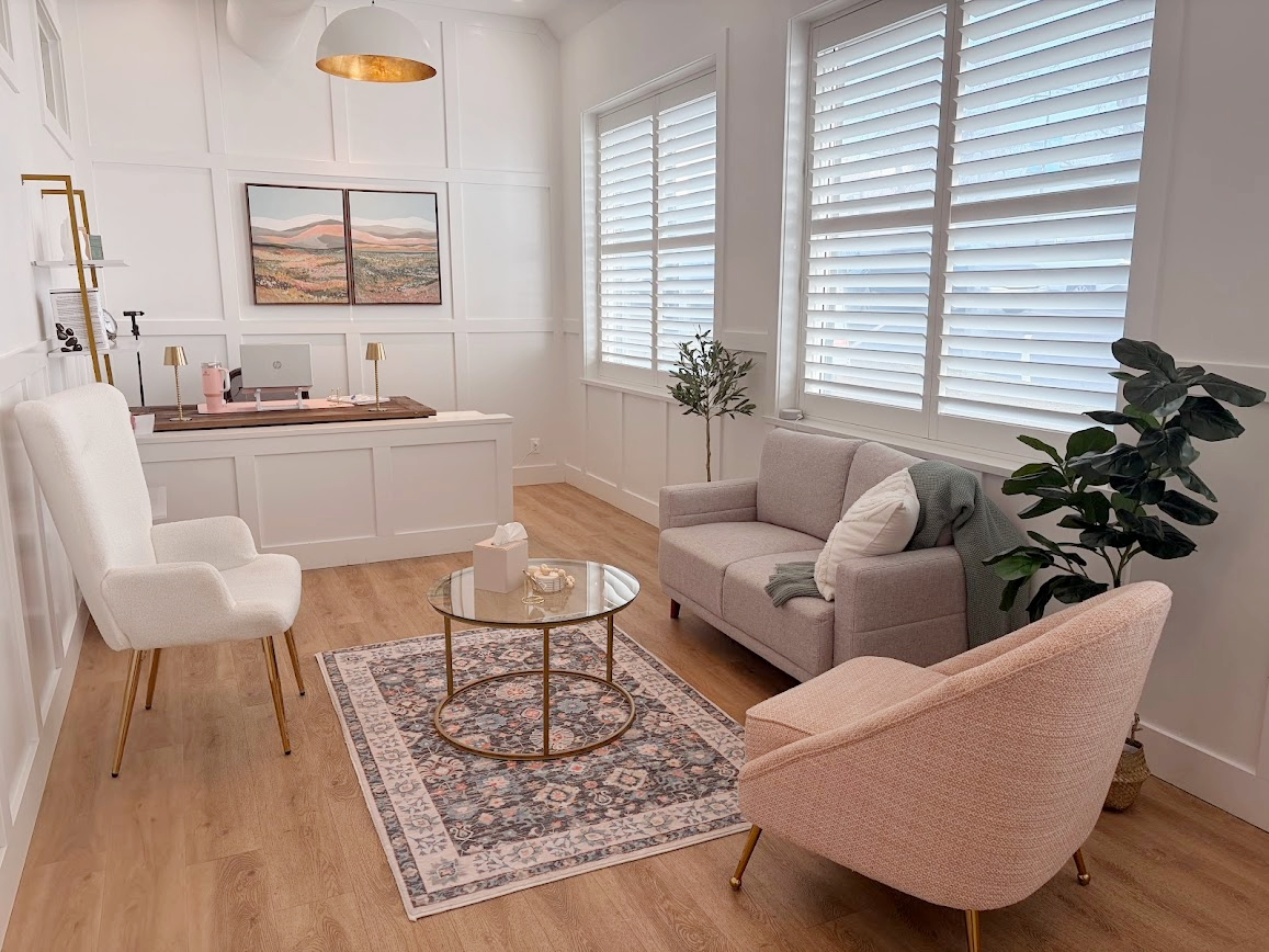 A cozy living room with white walls, large windows with shutters, a light gray loveseat, a pink armchair, and a white high-back chair. A round glass coffee table is placed on a patterned rug, with tissue box and decorative items on it. There is a small plant in the corner and framed artwork on the wall behind the desk area, which has a computer, lamp, and other office supplies.