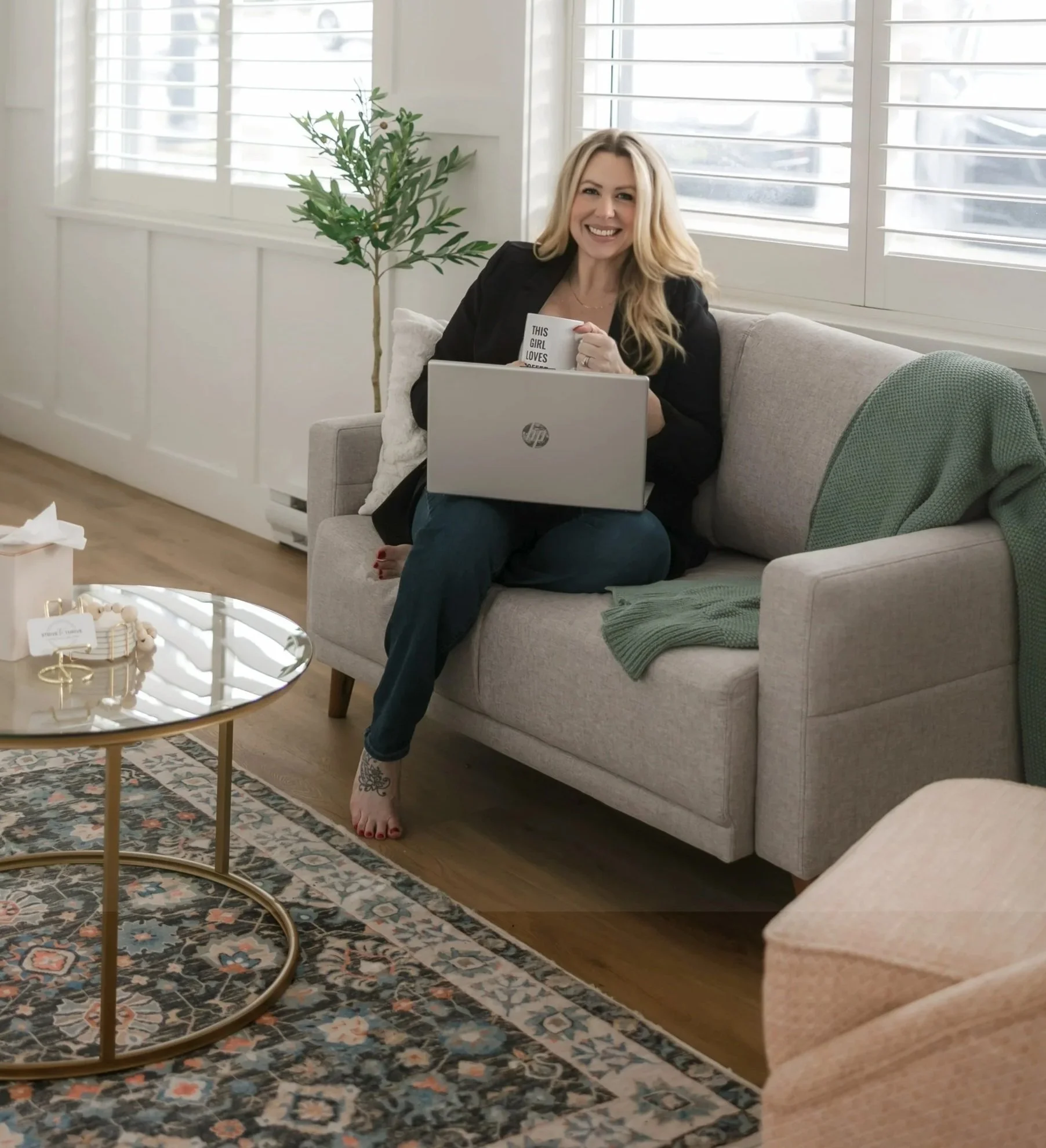 A woman with blonde hair sitting on a gray sofa, smiling, holding a mug that says "This girl loves...", with a laptop on her lap, in a bright living room with large windows, a green blanket, a potted plant, a gold coffee table, and a patterned rug.