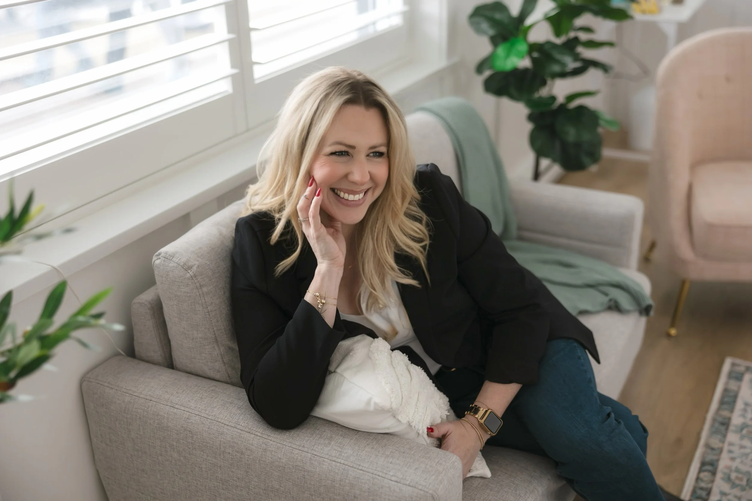 A woman with blond hair sitting on a sofa, smiling, with a window behind her and potted plants in a well-lit room.