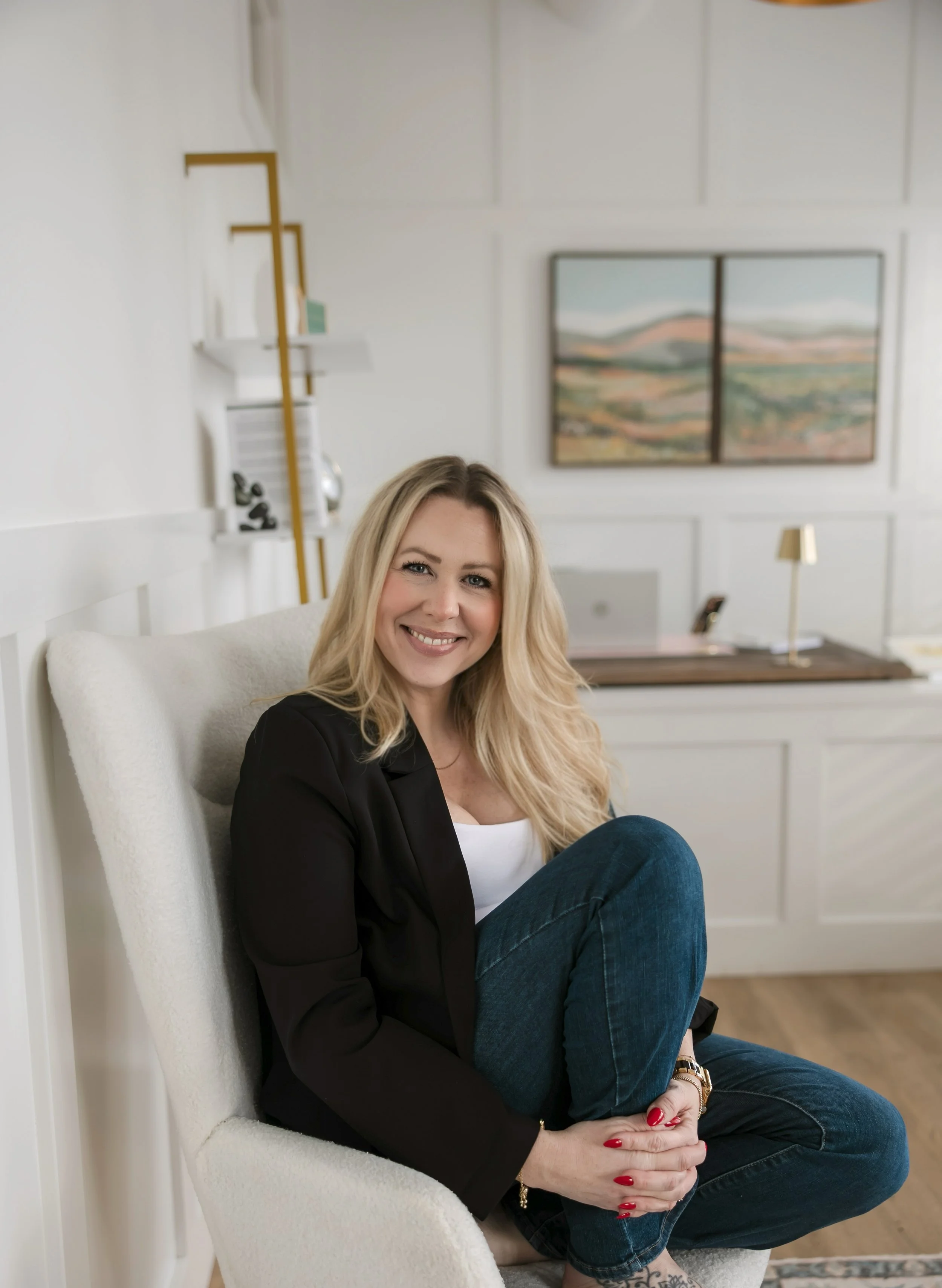 A woman with blonde hair smiling, sitting in a cream-colored armchair in a bright, modern room with white walls, artwork, and shelving.