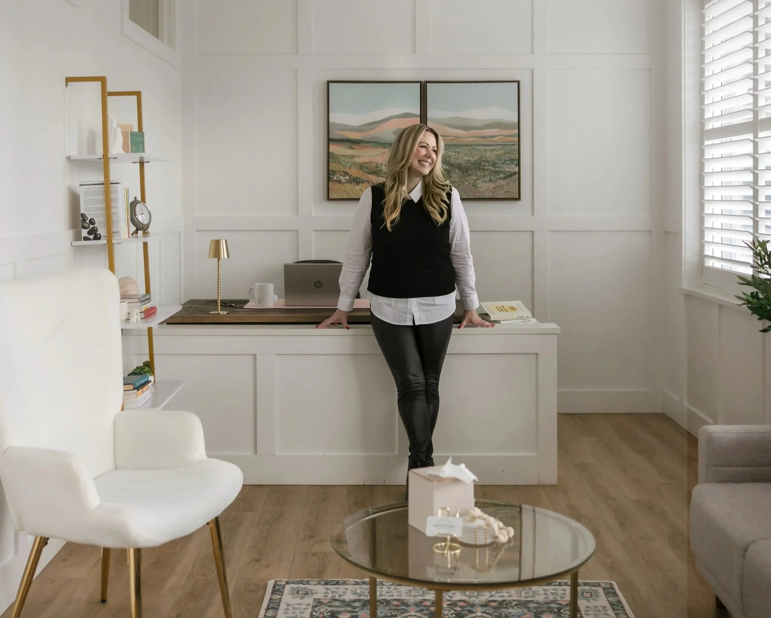 A woman standing behind a desk in a bright, modern living room with white walls and large windows with blinds, decorated with framed landscape art.