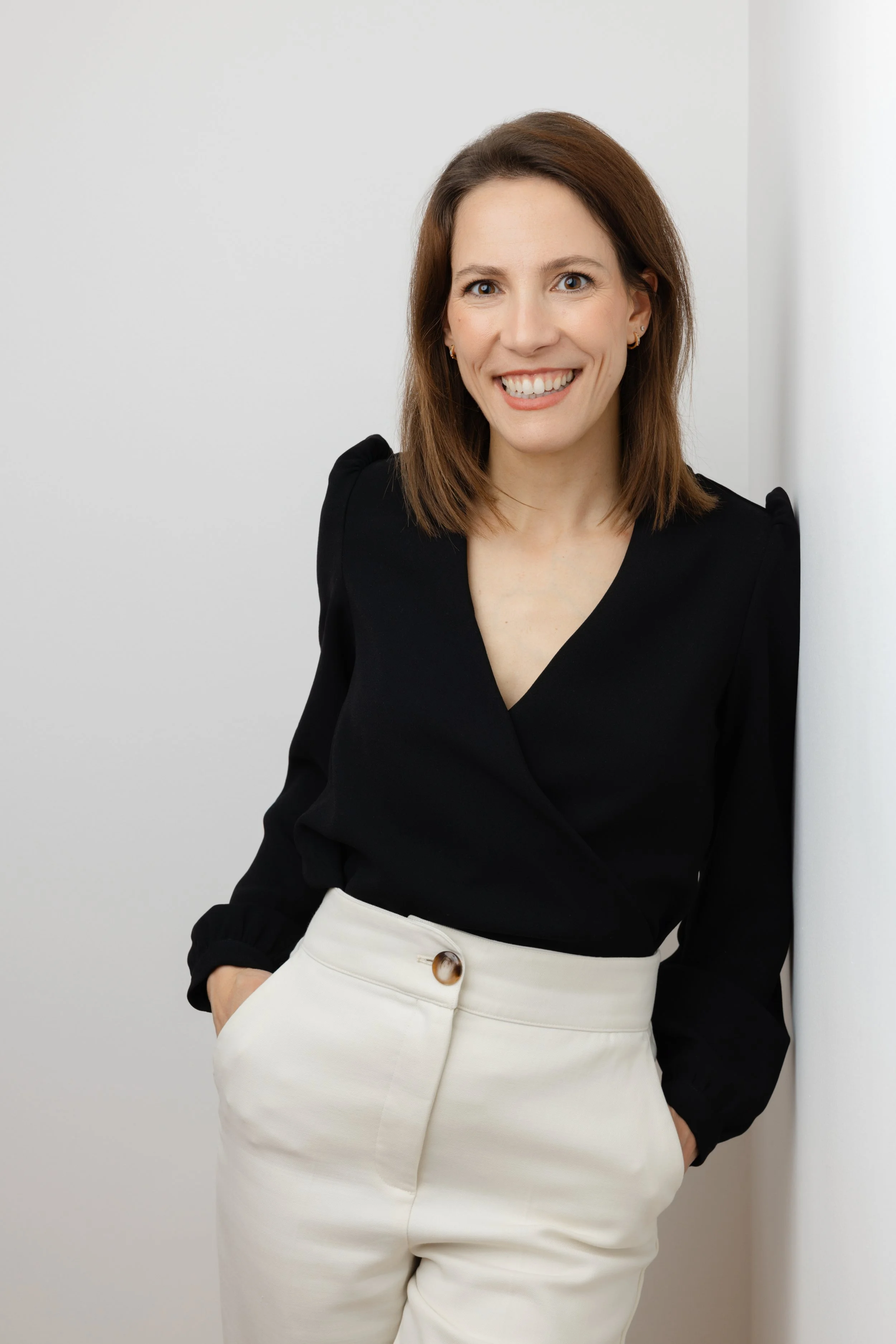 Femme souriante avec les mains dans les poches, portant une veste noire et un pantalon beige, posant contre un mur blanc.
