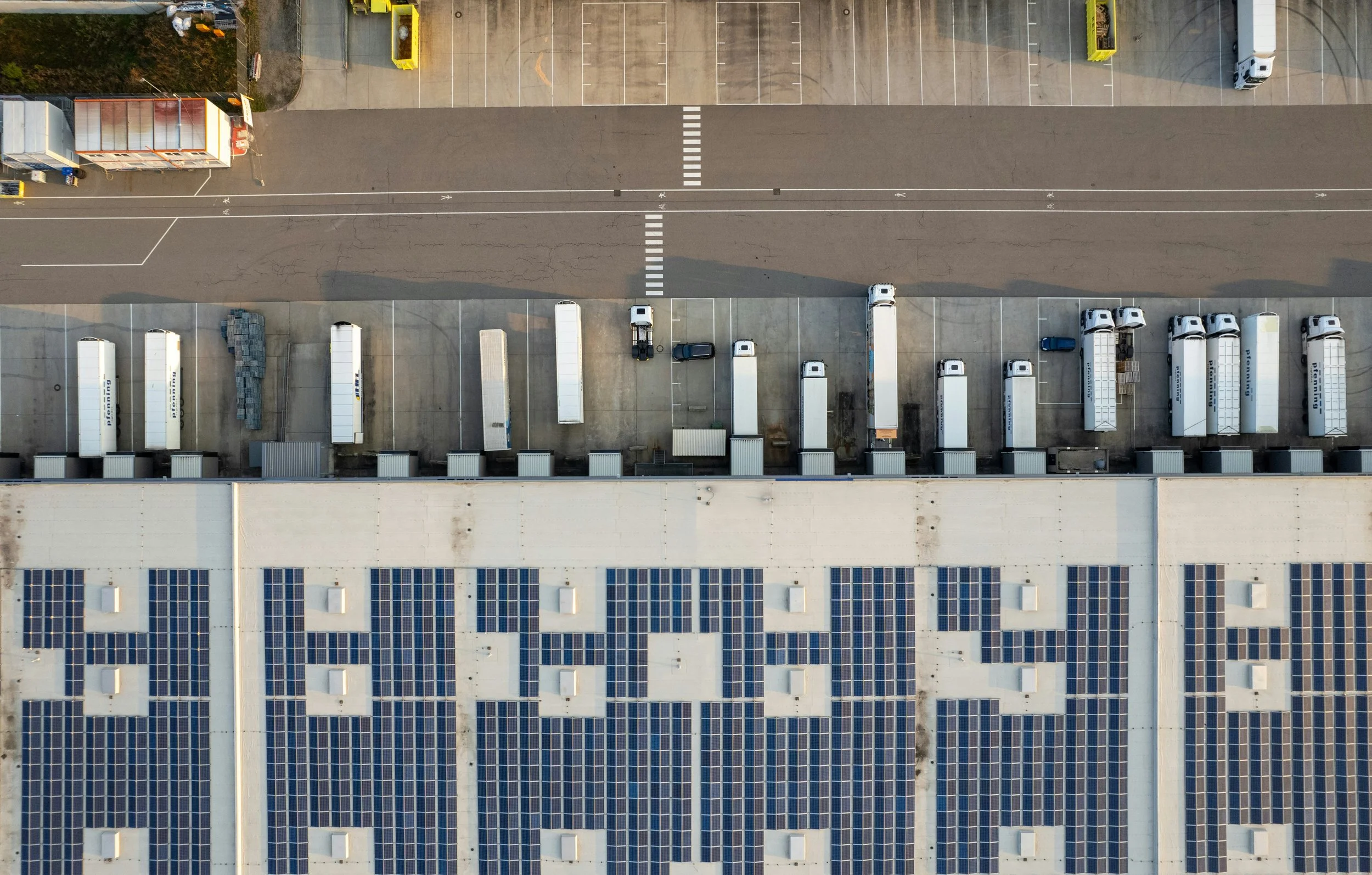 An aerial view of warehouse loading docks with trucks parked, solar panels on the roof, and a parking lot separated by a street.