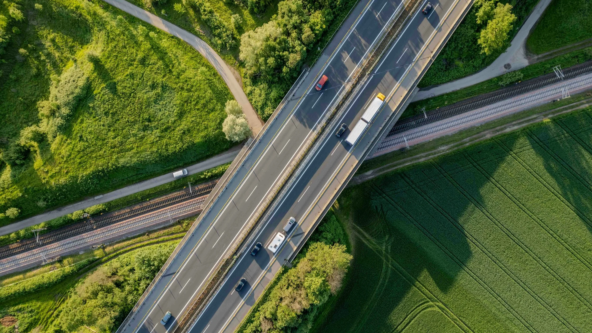 Aerial view of a highway bridge over green fields and trees, with cars and trucks traveling on it, and railroad tracks underneath.