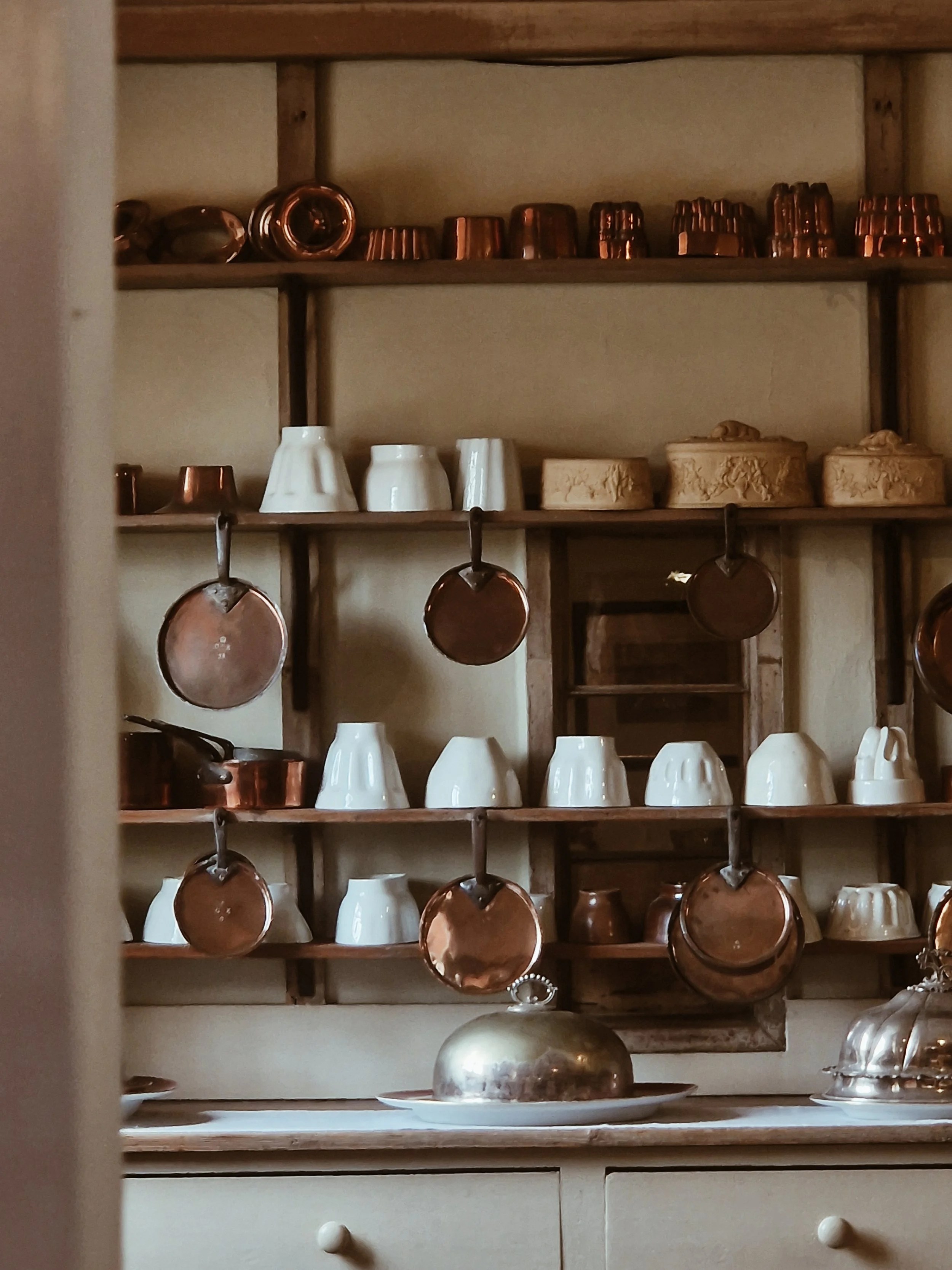 Rustic kitchen shelf with copper and white ceramics, hanging copper cups and pots, with a silver dome-shaped serving tray on a countertop.